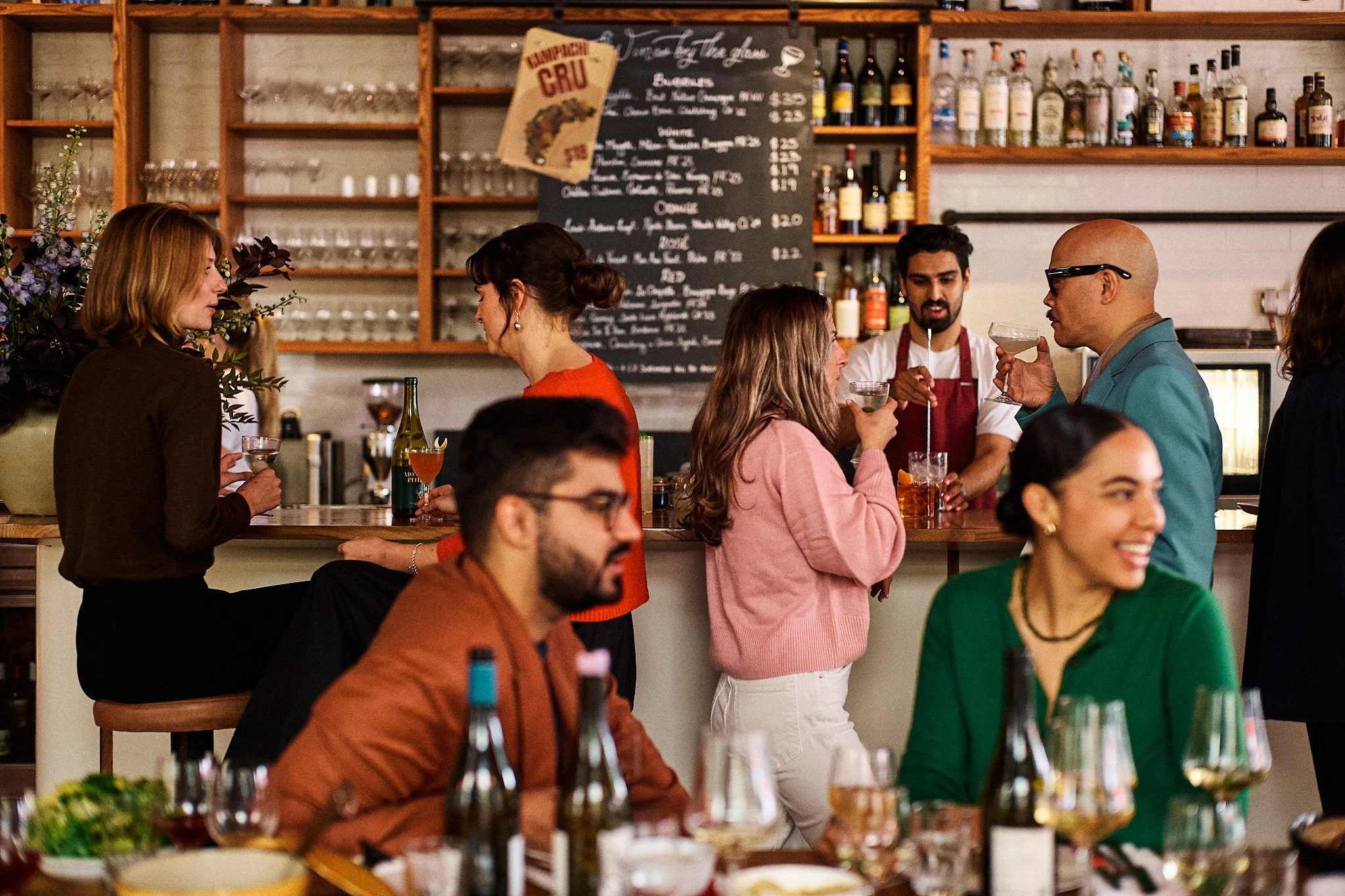 People socializing at a bar, some holding drinks and engaging in conversations, with a menu on a chalkboard behind the bar.