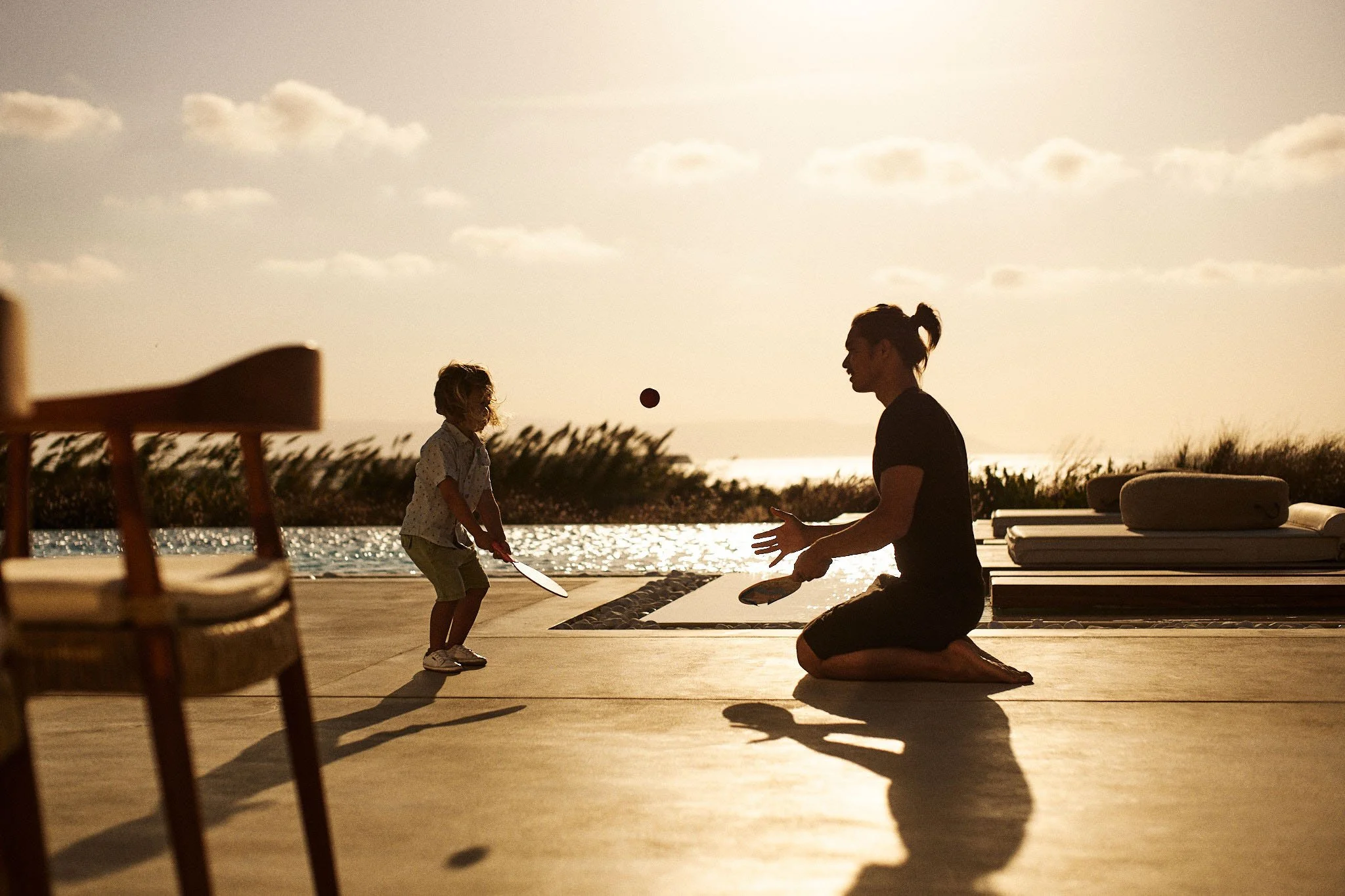 A silhouette of a woman and a child playing with a ball and a racket outdoors during sunset, near a pool with lounge chairs and a beach with tall grass in the background.