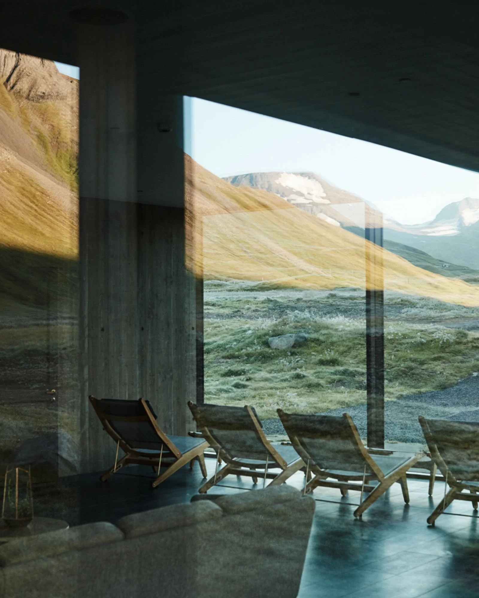 Interior view of a room with large floor-to-ceiling windows showing a mountainous landscape with grassy slopes and snow-capped peaks; there are several wooden lounge chairs facing the window.
