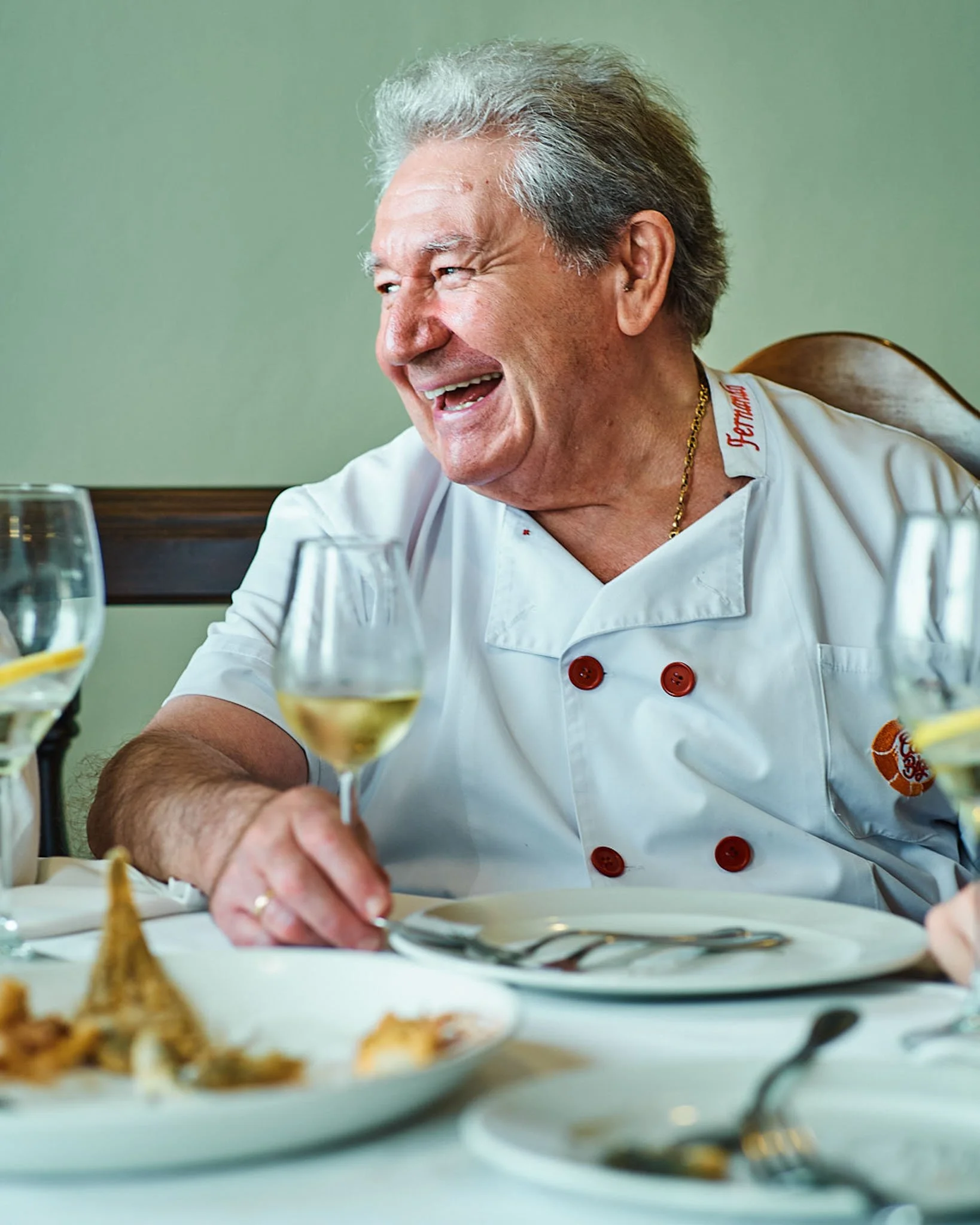 An elderly man in a chef's uniform sitting at a table with wine glasses, smiling and enjoying a meal.