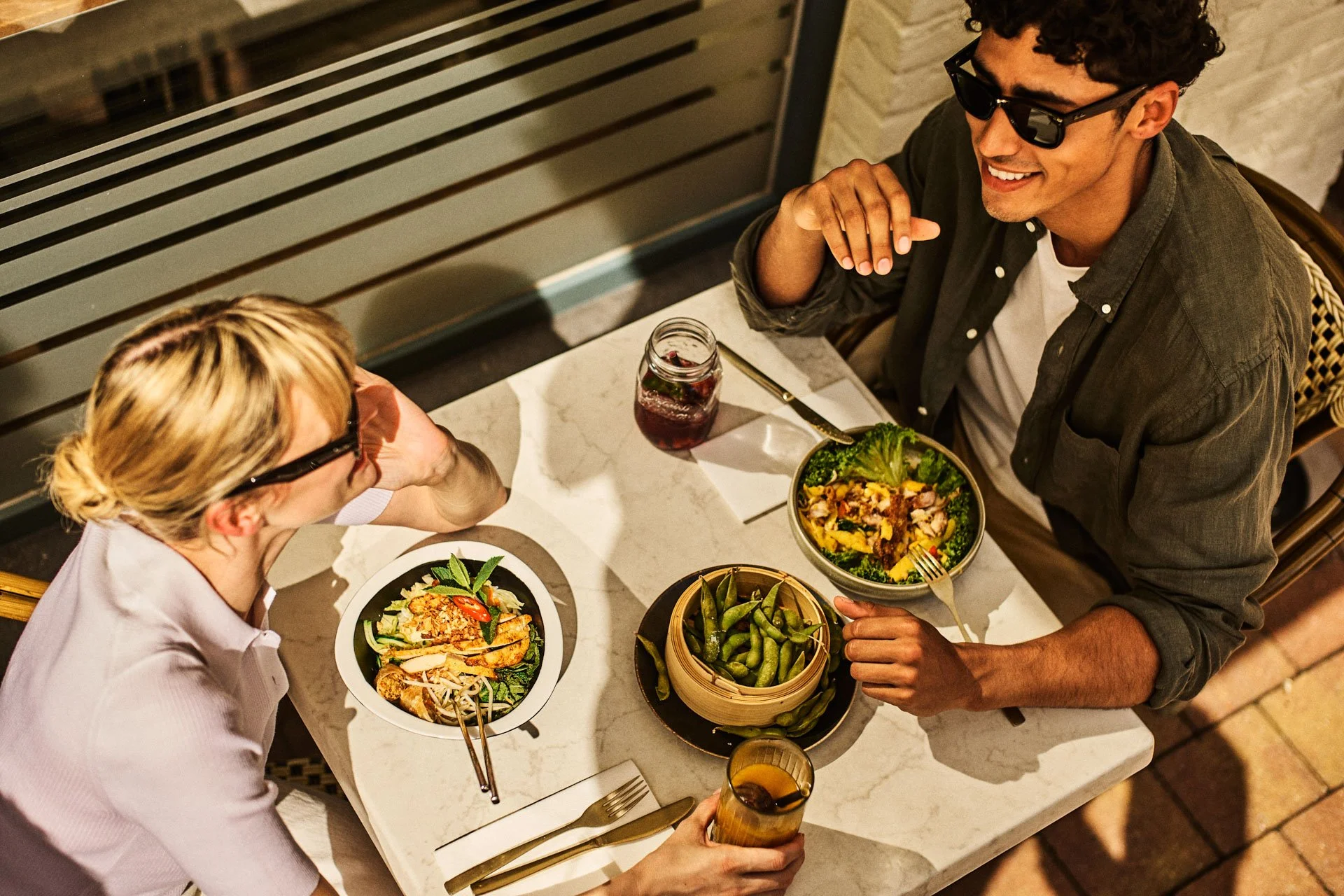 A man and woman sitting at a table enjoying a meal outdoors. The woman has blonde hair tied back, wearing glasses and a white shirt, and the man has curly dark hair, glasses, a grey jacket, and is smiling. They have salads, edamame, and drinks on the