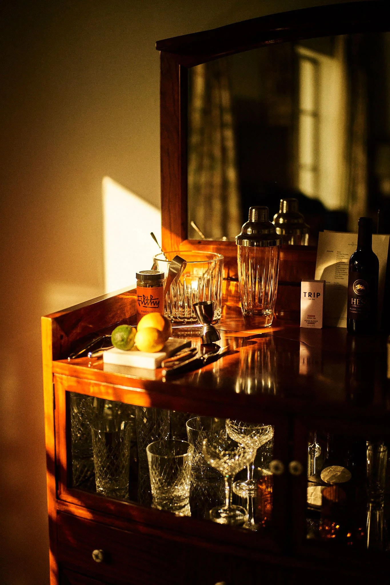 A wooden bar cabinet with glassware and bottles, illuminated by warm sunlight