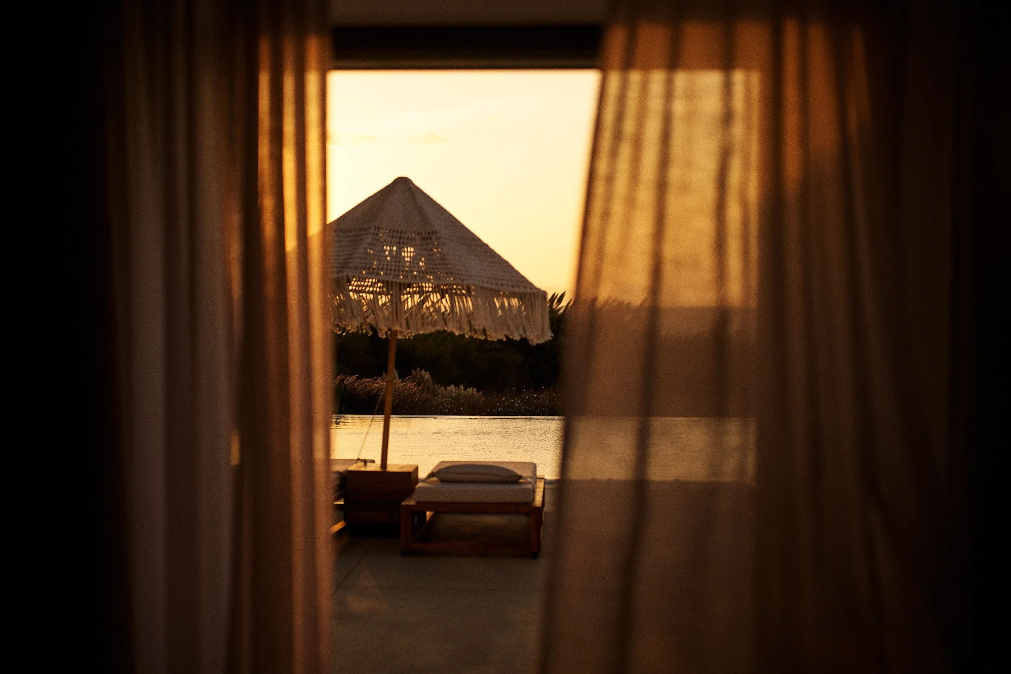 Beach scene viewed through window with beige curtains, showing a parasol and lounge chair near water at sunset.