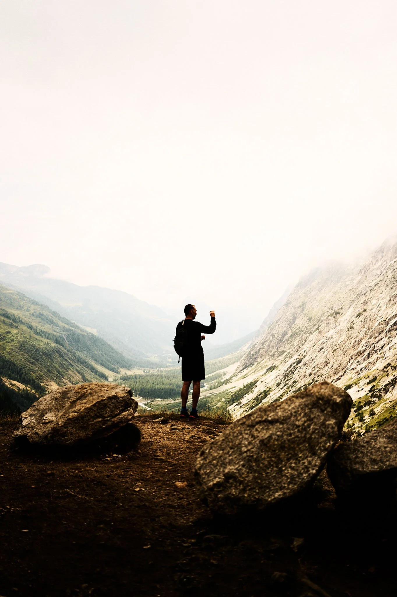 Man standing on a rocky trail in a mountainous area, raising his fist in the air, with large boulders and a deep valley in the background.
