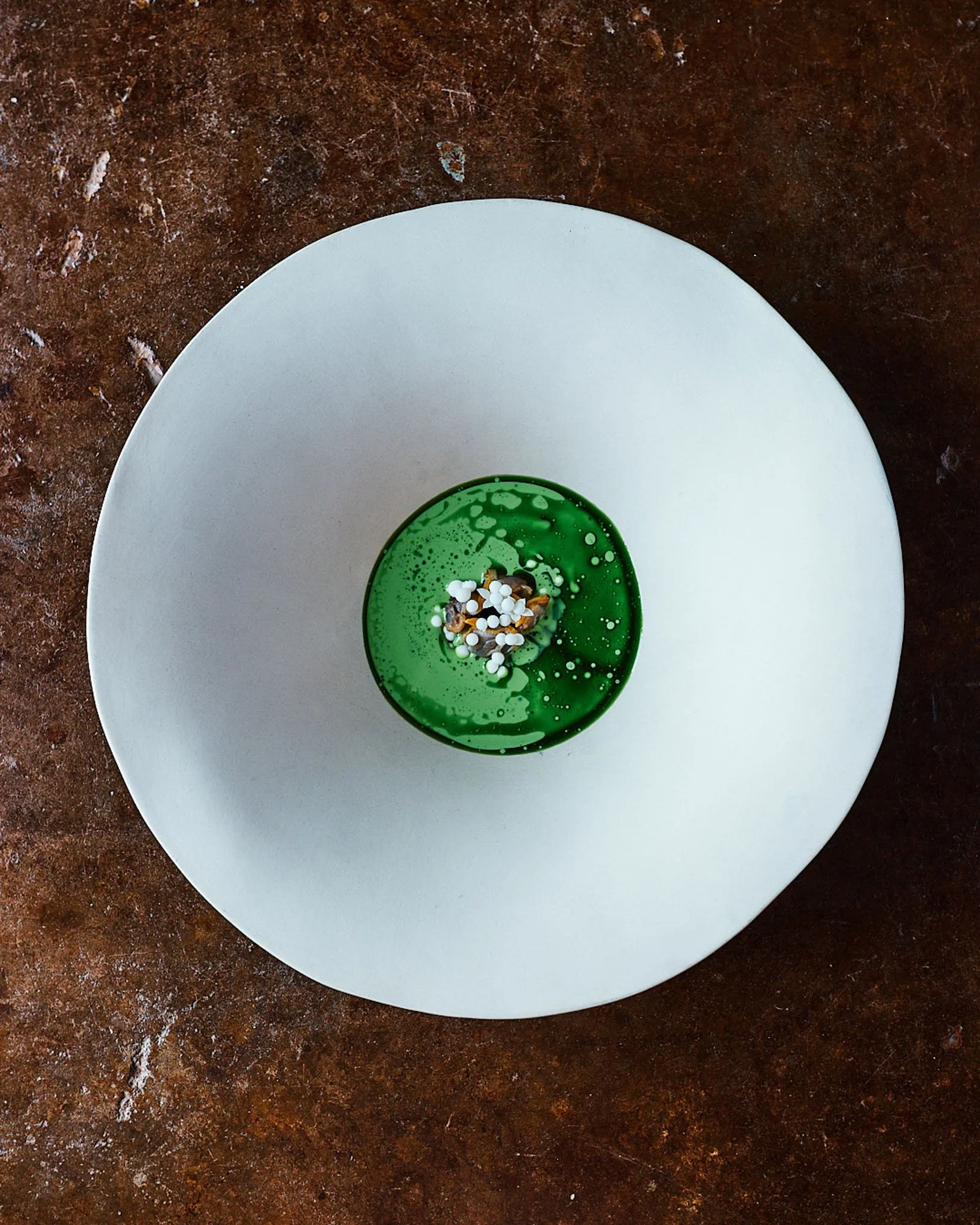 A white plate with a small green glass container filled with a green liquid, garnished with small white and dark sprinkles, placed on a textured brown surface.