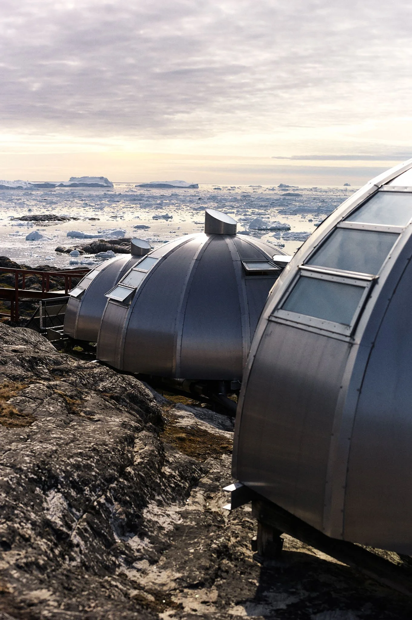 Research observatories with metallic domes on a rocky landscape overlooking an icy seascape with floating icebergs under a cloudy sky.