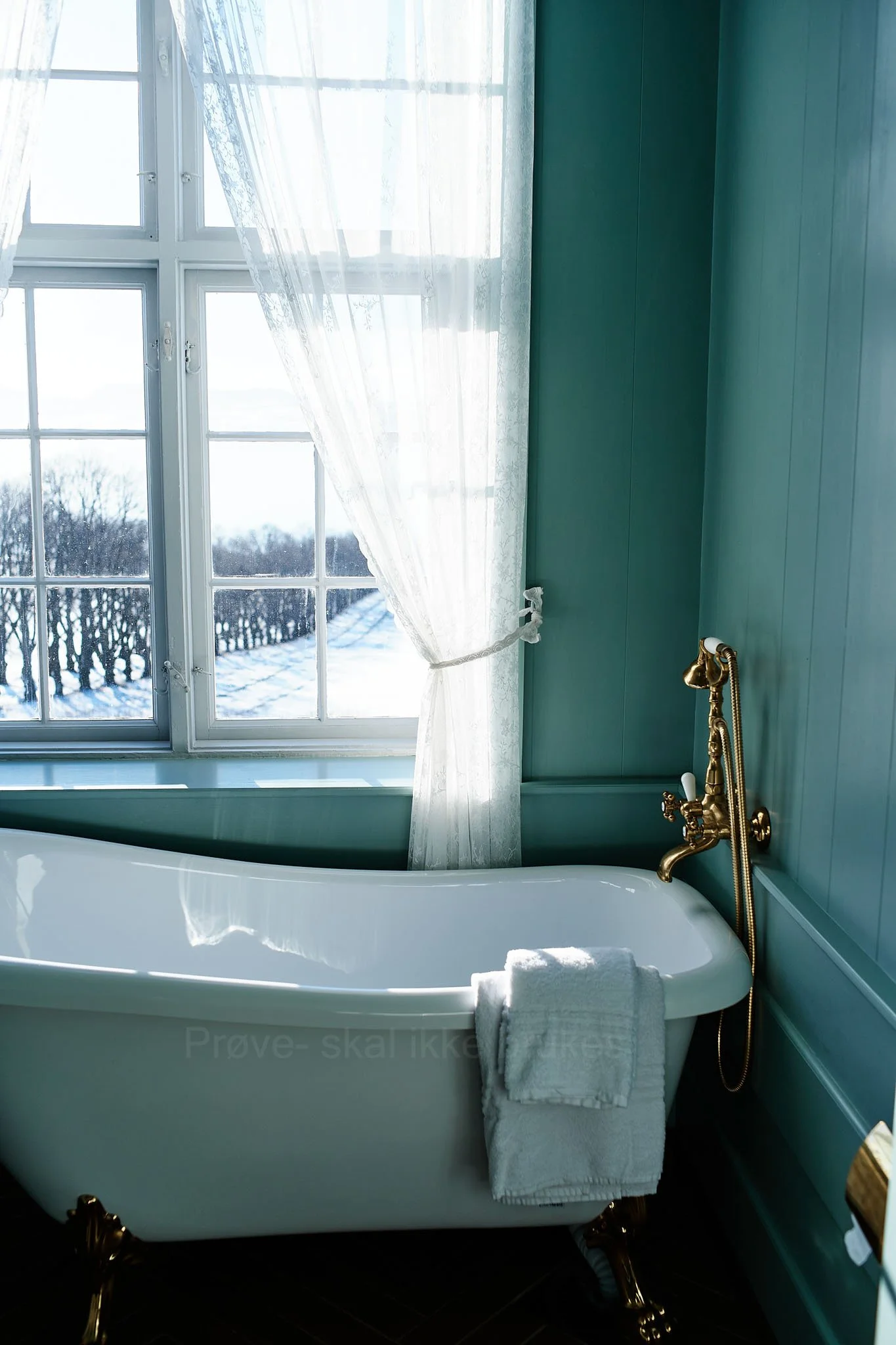 Vintage white bathtub with gold fixtures next to a window with lace curtains, overlooking a snowy outdoor scene.