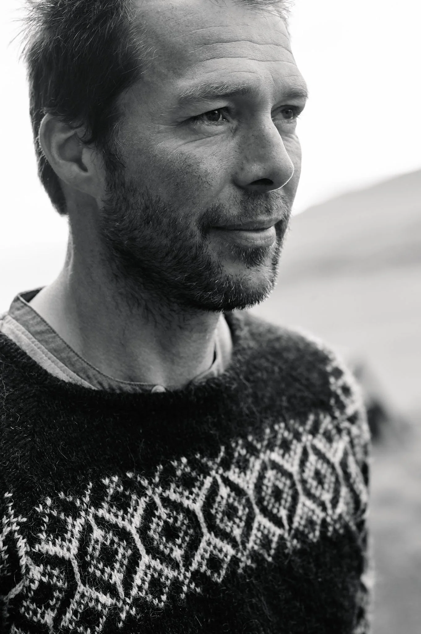 A black and white image of a middle-aged man with a beard and short hair, wearing a patterned sweater and looking thoughtfully into the distance outdoors.