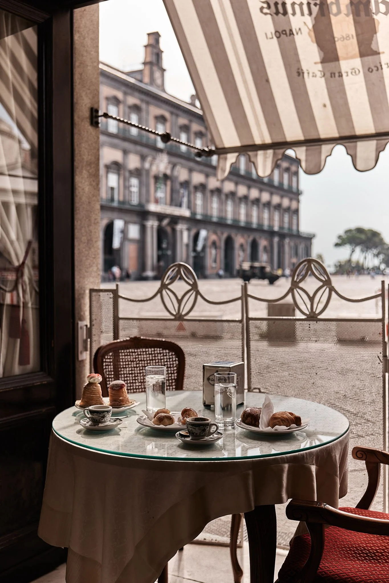 A table set with pastries, teacups, and glasses of water in a cozy cafe with a view of a European-style building outside.