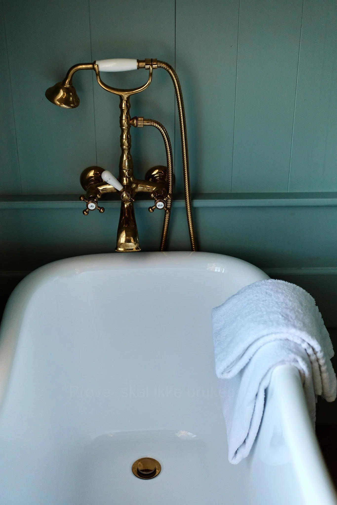 A vintage-style white bathtub with a white towel draped over the side, situated against a green paneled wall with a brass faucet and shower head.