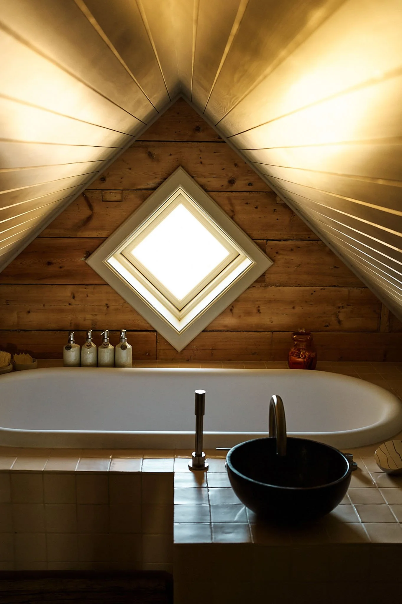 A bathroom with a bathtub beneath a skylight, wooden walls, and a black bowl sink in front.