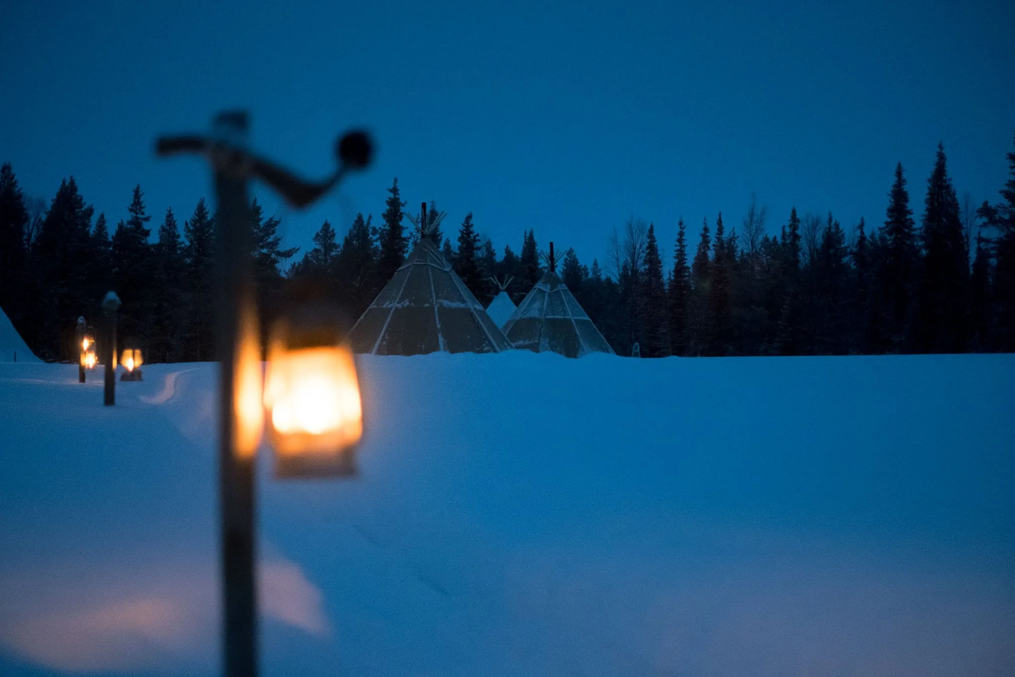 Snow-covered landscape at dusk with illuminated lanterns leading to teepees and a group of trees in the background.