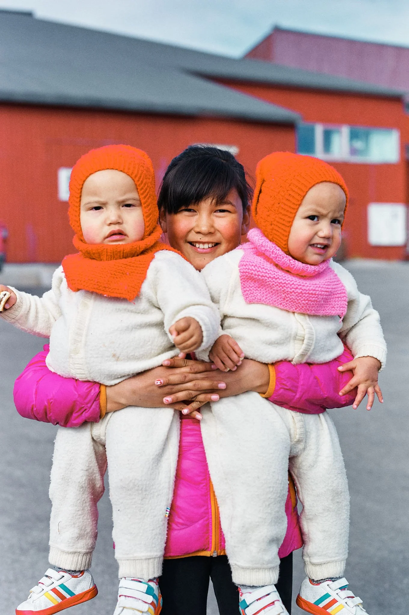A woman holding two young children on her shoulders outdoors, all wearing warm clothing with orange knit hats, in front of a red building.