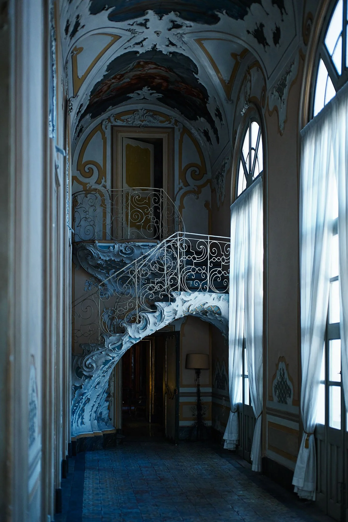 Interior of an ornate room with blue and white decorative molding, large arched windows with sheer curtains, and a white wrought iron staircase with intricate swirls and curling designs.