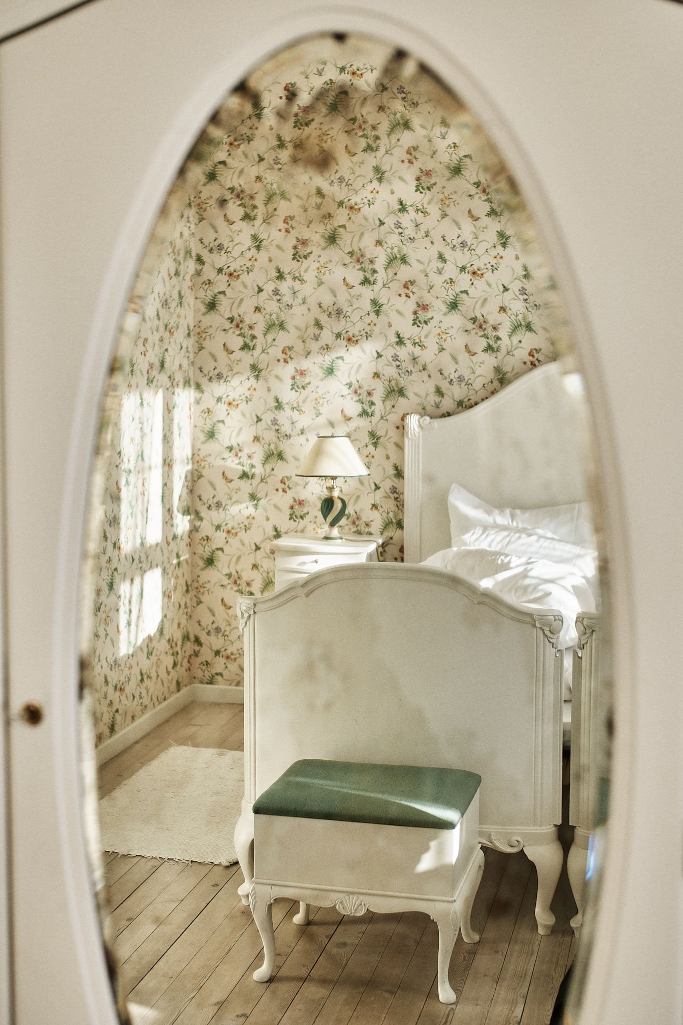 A bedroom with floral wallpaper, a white bed with pillows, a white nightstand with a lamp, and a small vintage-style bench with a green cushion, reflected in an oval mirror.