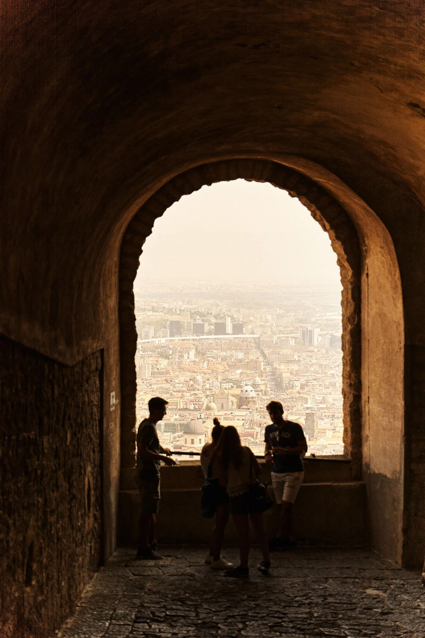 Four people standing inside a stone tower with a large arched window overlooking a cityscape, likely during sunset.