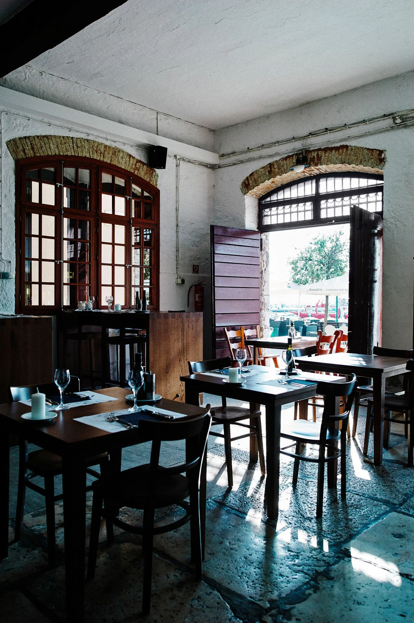 Interior of a restaurant with wooden tables and chairs, set with wine glasses, candles, and utensils. Sunlight streams through an open arched doorway revealing outdoor seating and a view of the street.