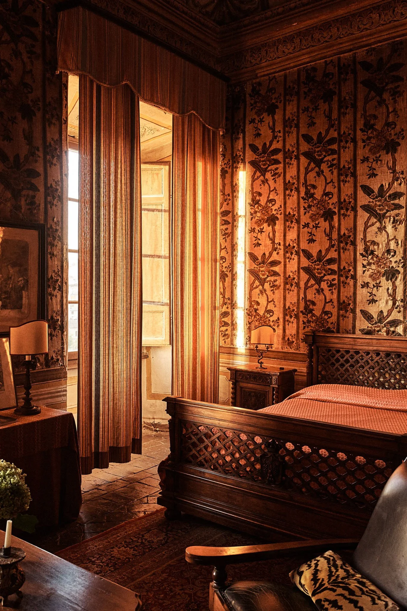 A bedroom with wooden furniture, floral patterned walls, and striped curtains in warm tones, illuminated by sunlight through open shutters.