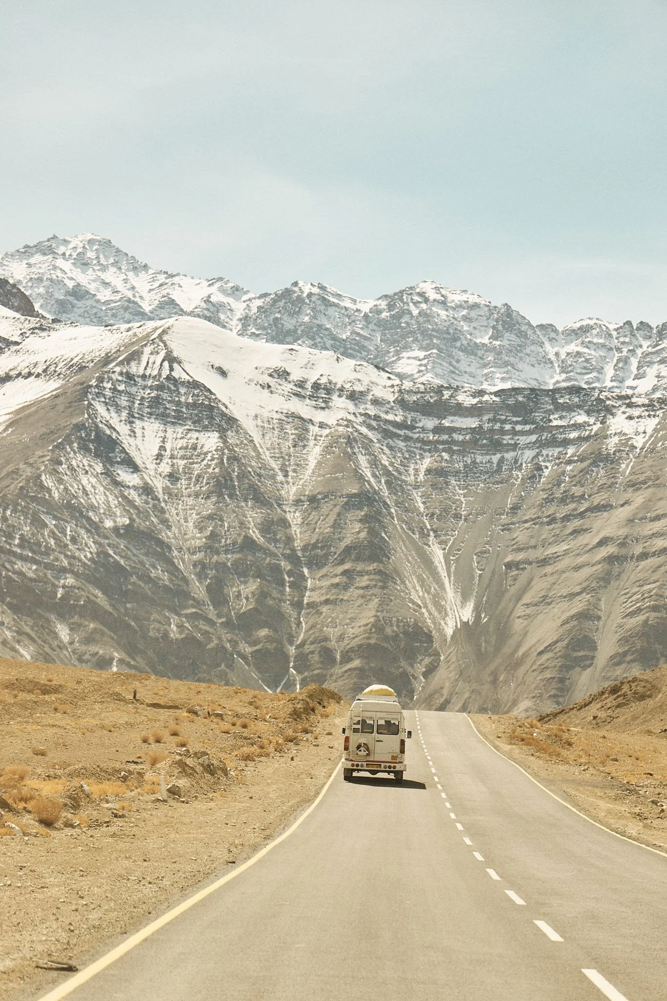A camper van traveling on a winding mountain road surrounded by barren landscape and snow-capped mountains in the distance.