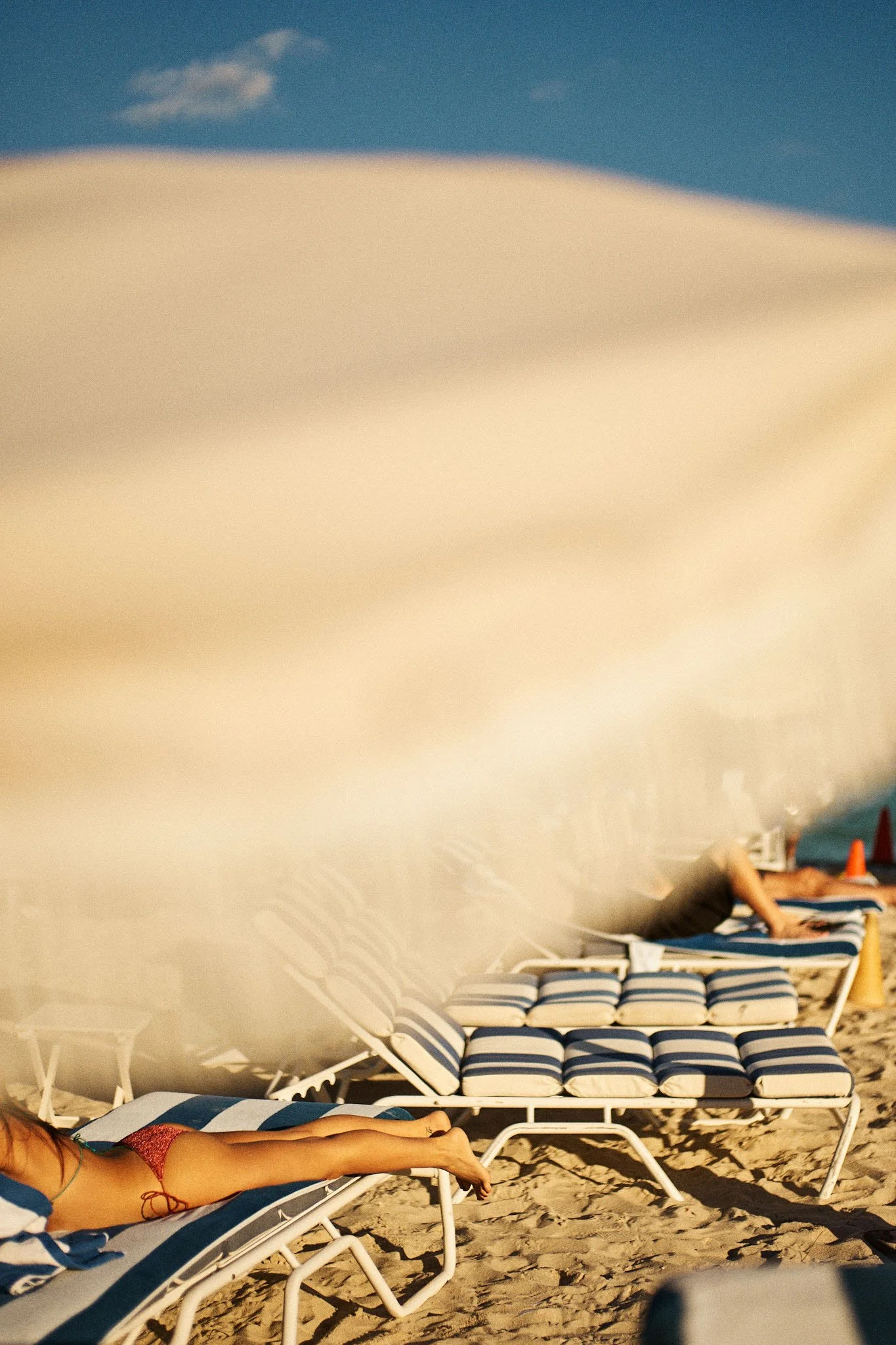 A person is lying on a beach lounge chair with striped cushions, partially obscured by a large white beach umbrella. There are more lounge chairs and another person in the background, with a beach, sand, and sky visible.