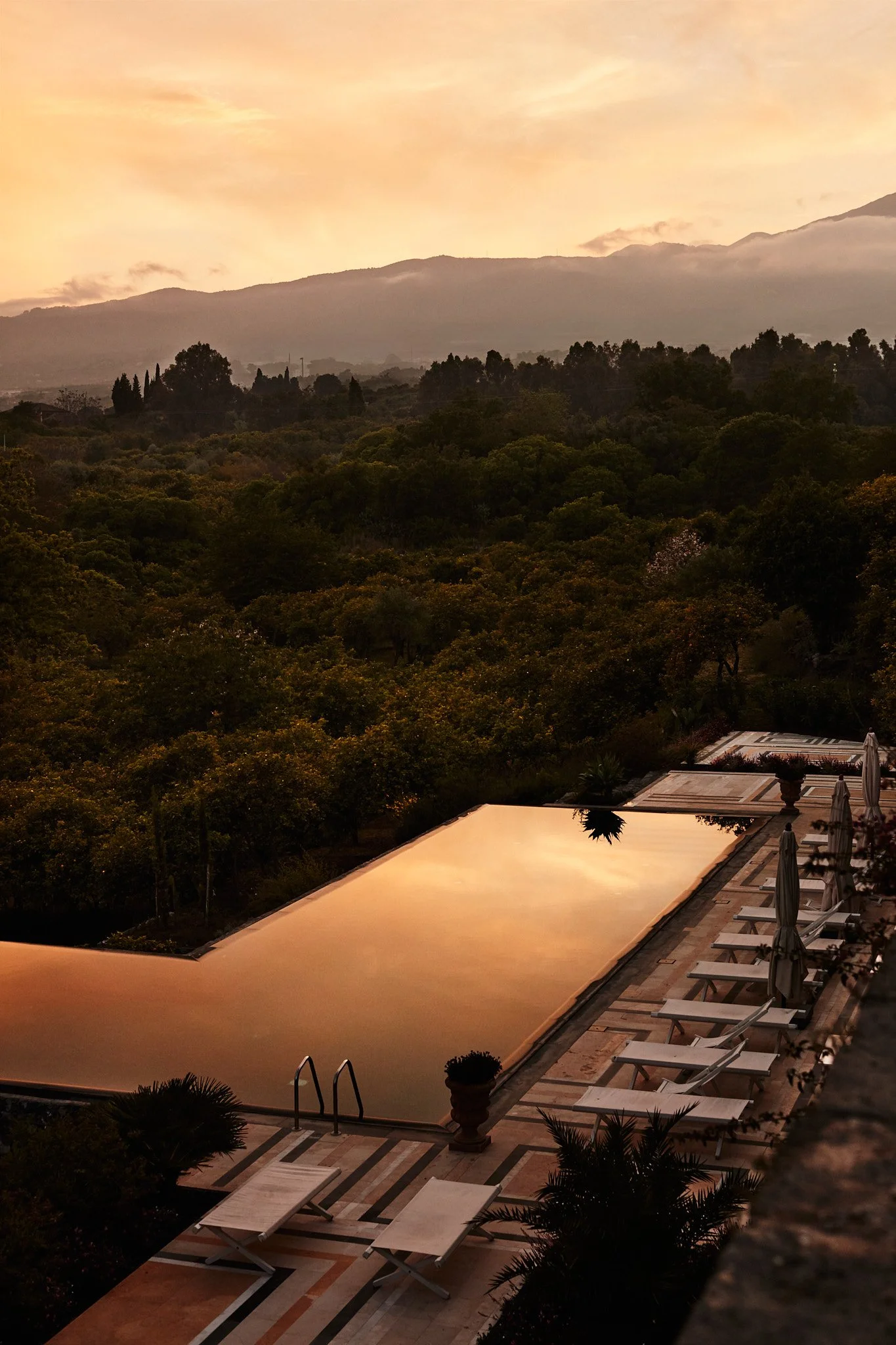 A scenic view at sunset with an infinity pool reflecting the sky, surrounded by lounge chairs and umbrellas overlooking a lush green forest and mountains in the distance.