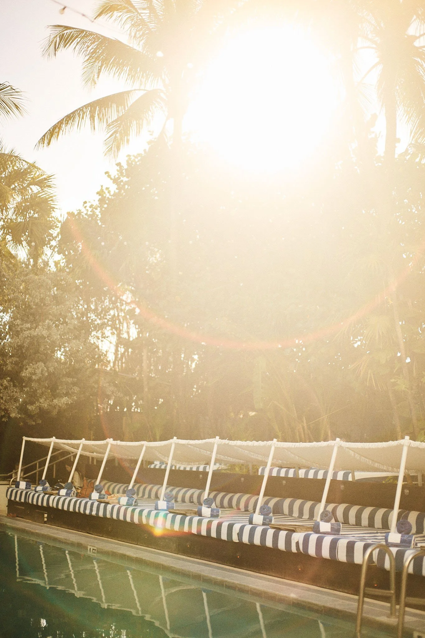 A poolside lounge area with striped cushions and a canopy, set against a backdrop of tropical palm trees and a bright sun.