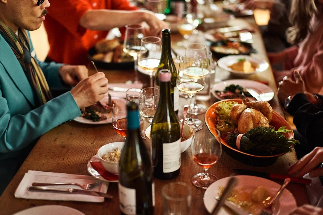 People dining at a table with wine bottles, glasses of wine, plates of food, and a roasted chicken.