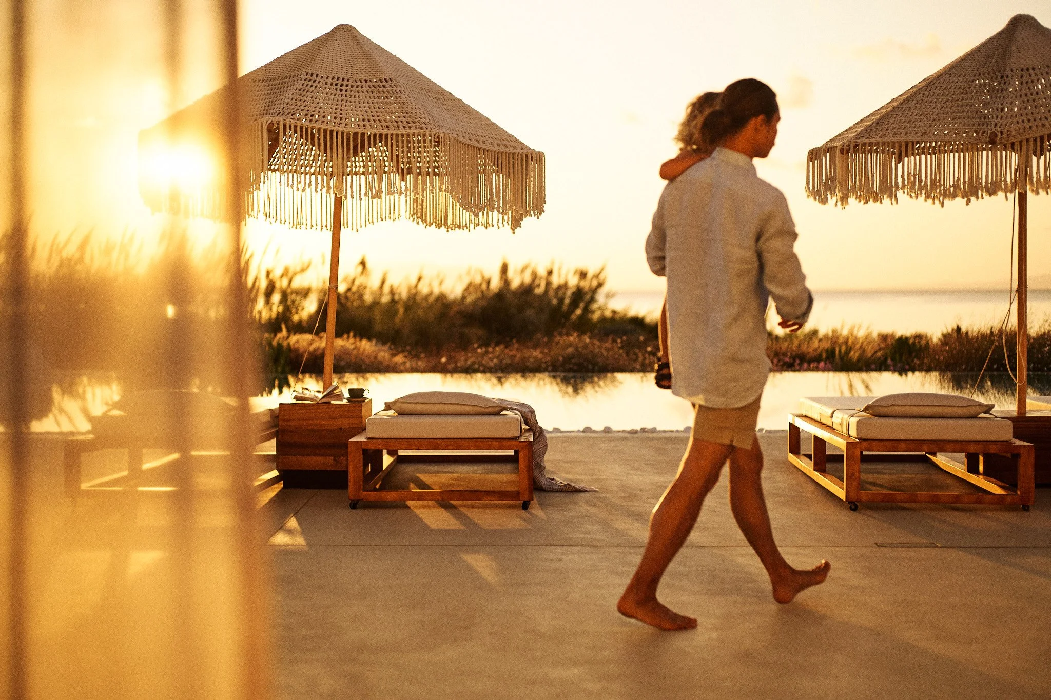 A woman walking barefoot on a pool deck during sunset, with outdoor bed and large umbrellas in the background.
