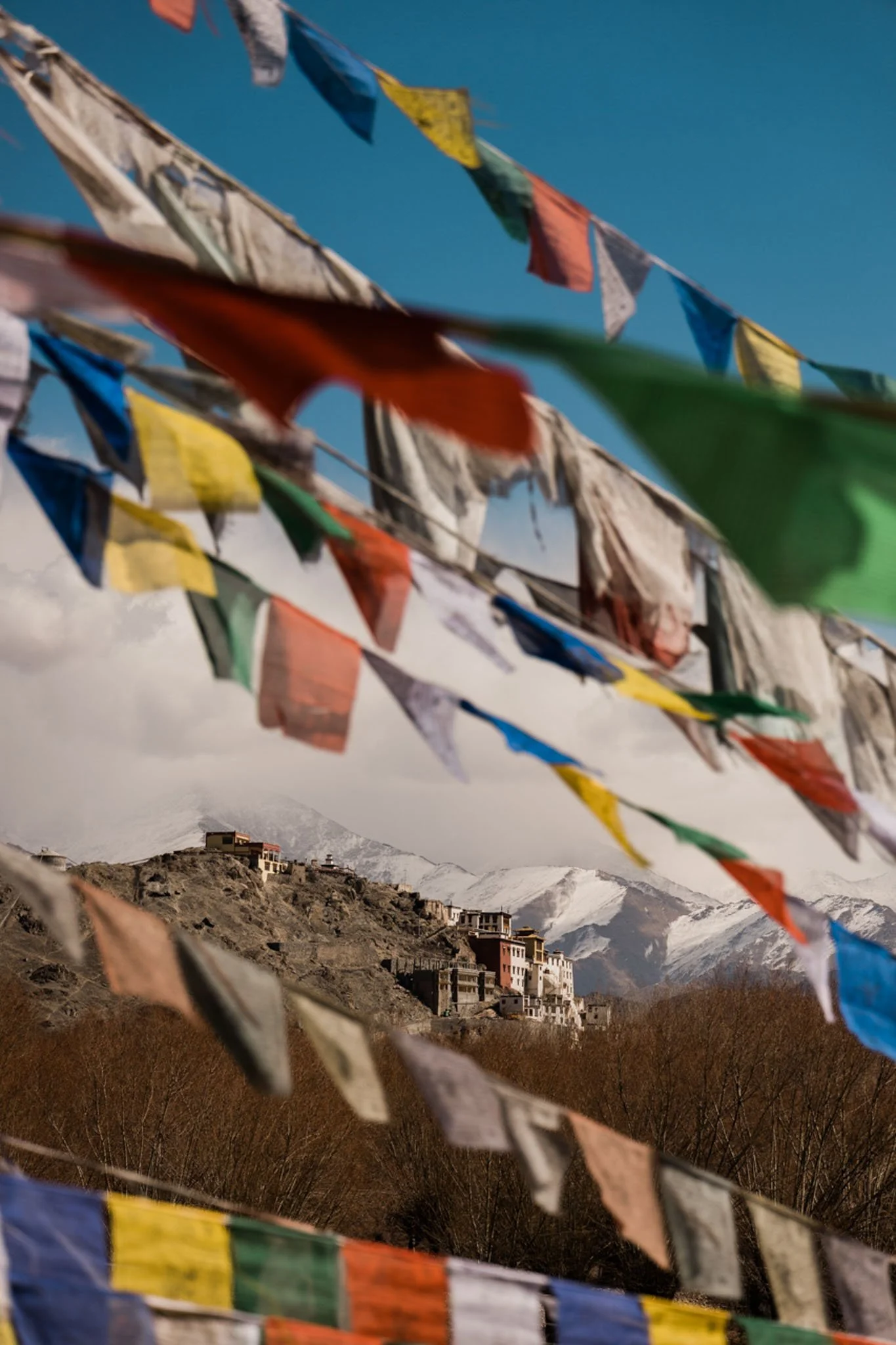 Colorful prayer flags in the foreground with snowy mountain peaks and buildings on a hillside in the background.