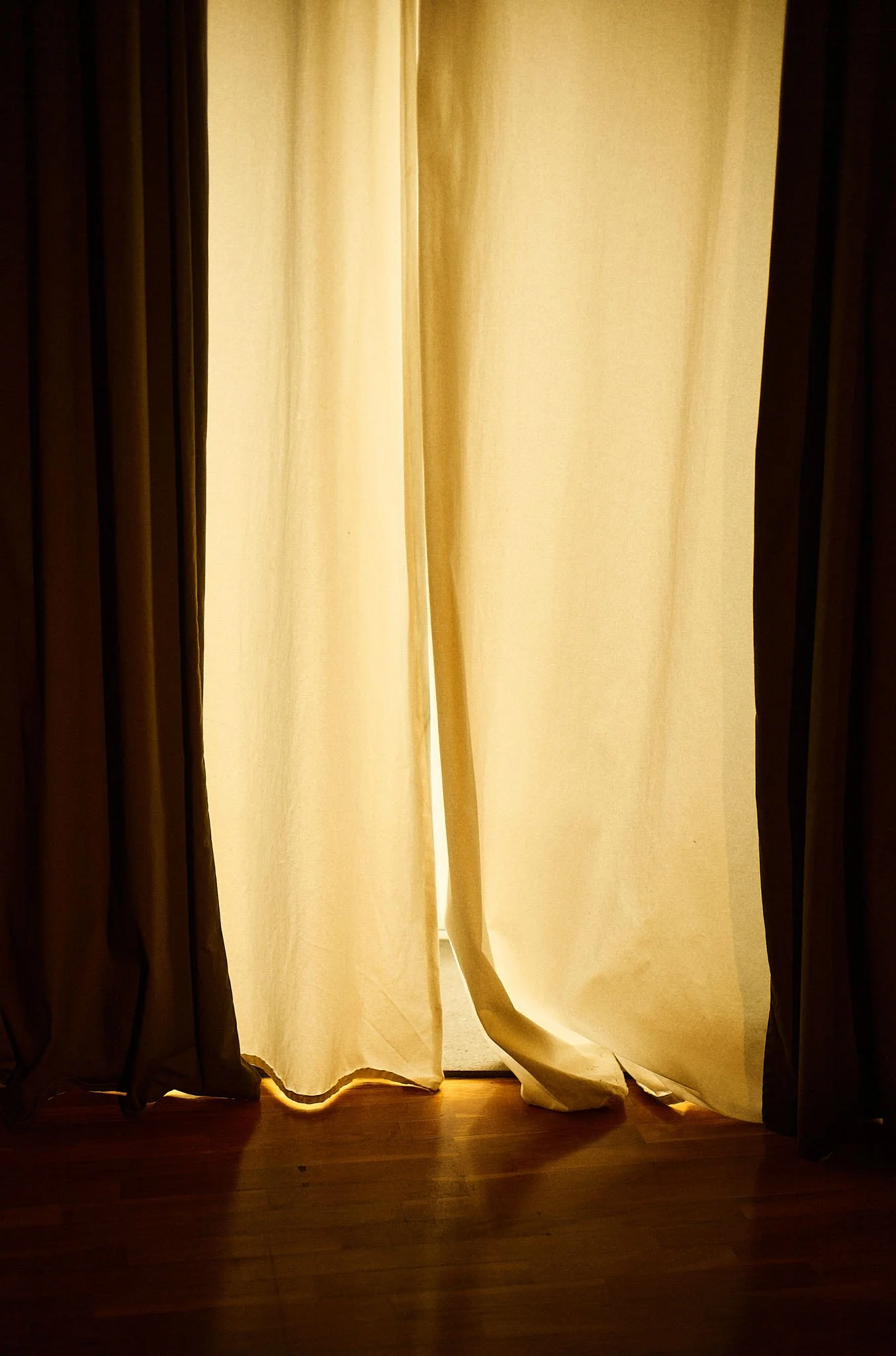 Closed yellow curtains with some light shining through at the bottom, covering a doorway or window, with a wooden floor visible at the bottom.