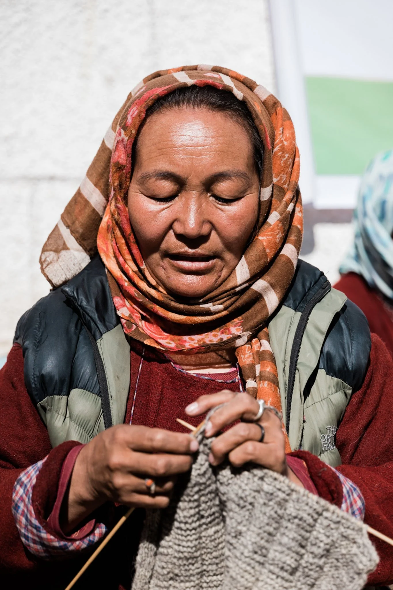 A woman with a headscarf, wearing a red sweater and a gray vest, knitting with gray yarn and knitting needles.