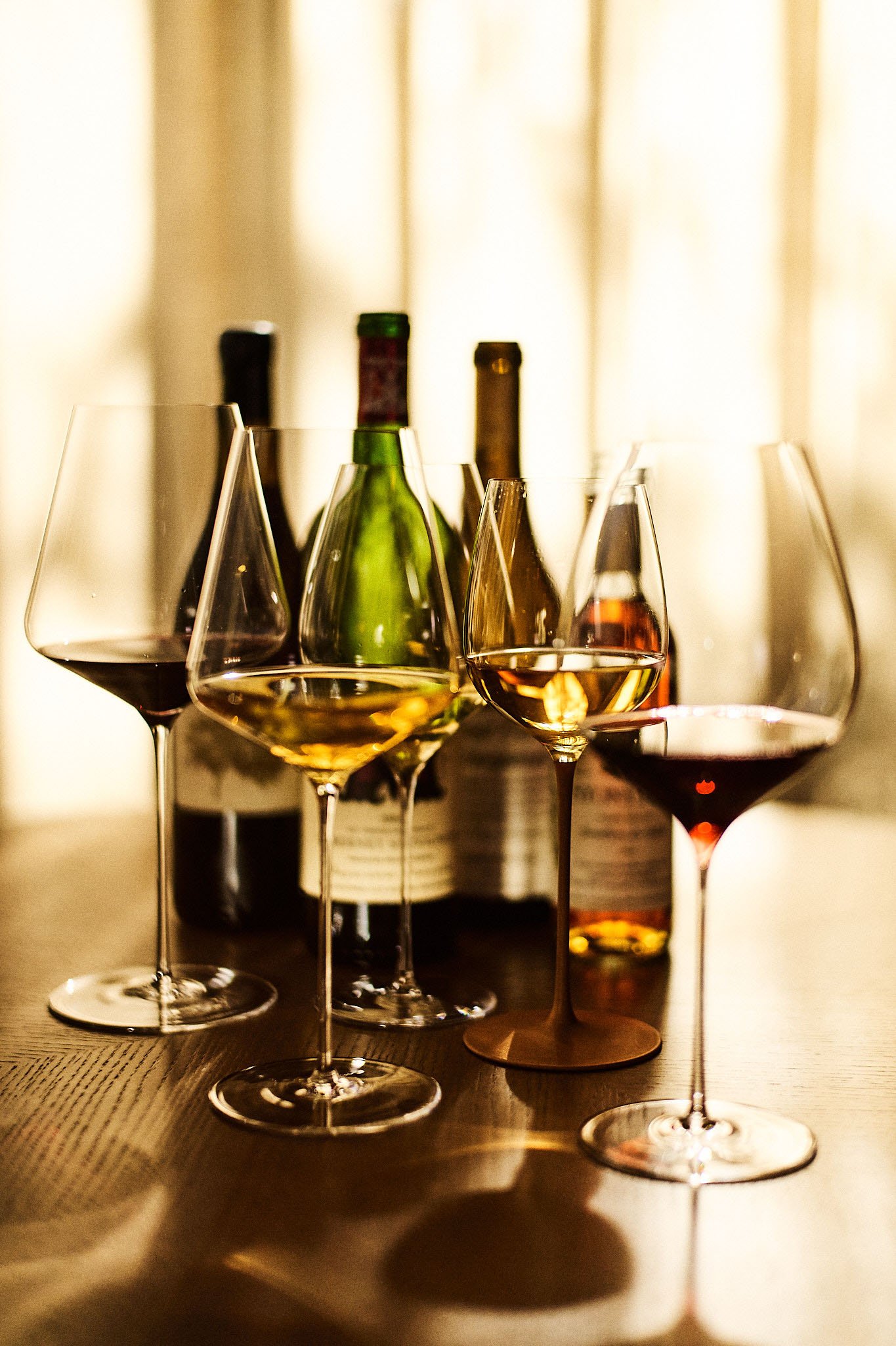 Several wine bottles and five filled wine glasses on a wooden table in front of a light-colored curtain.