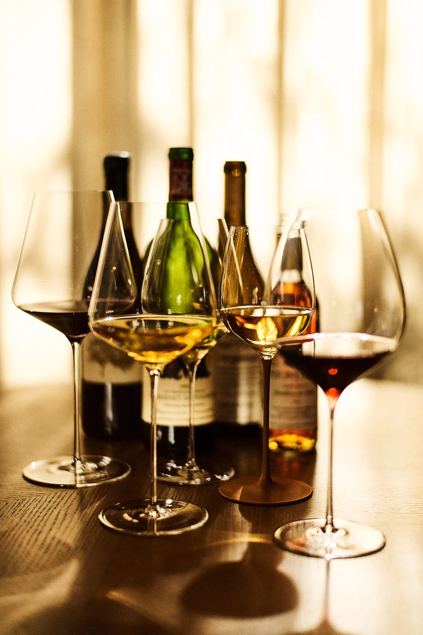 Several wine glasses filled with red and white wine on a wooden table, with wine bottles in the background near a window with curtains.