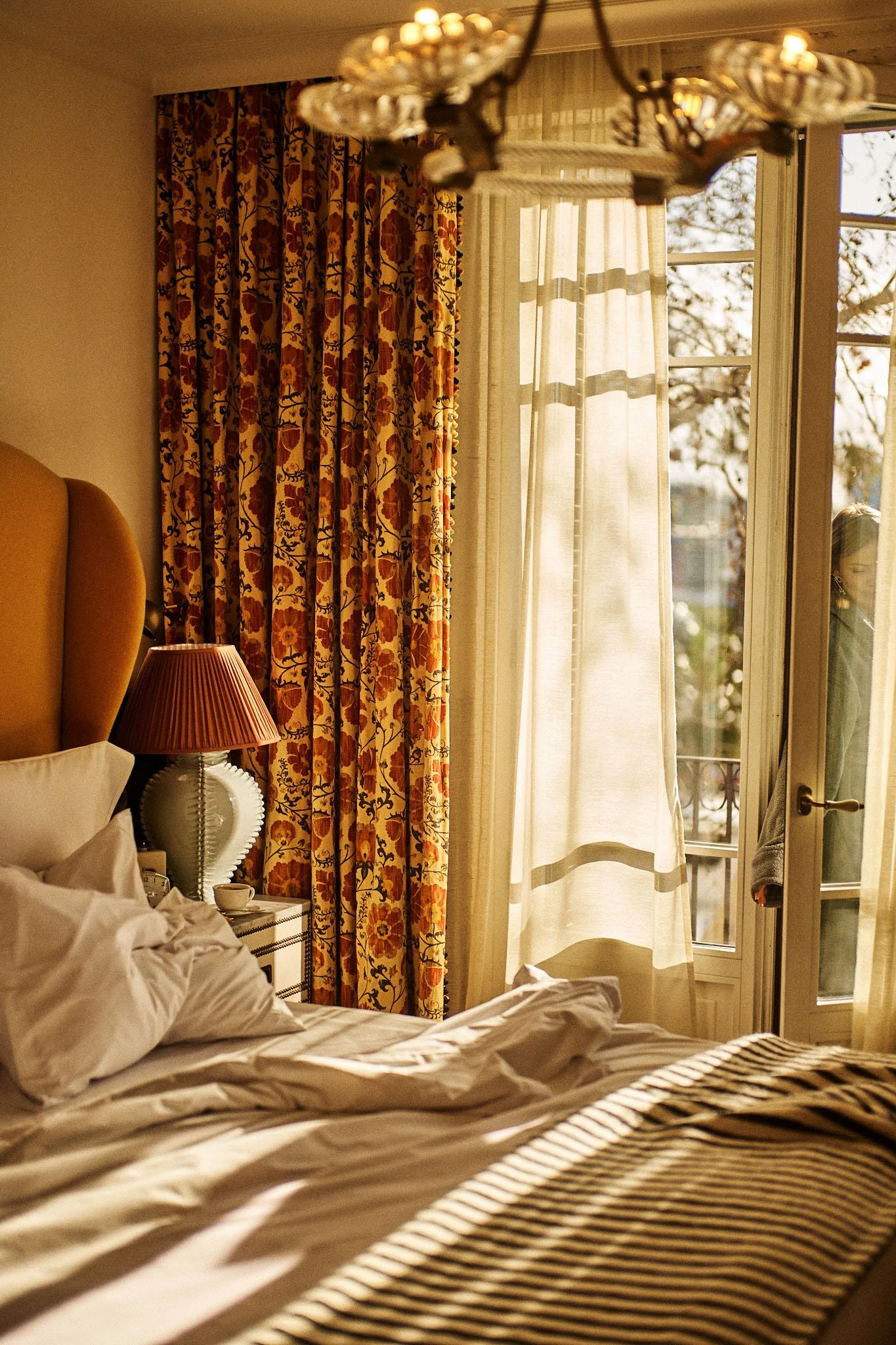 A cozy hotel room with a bed, a bedside table with a lamp, floral curtains, and a woman standing outside on a balcony.