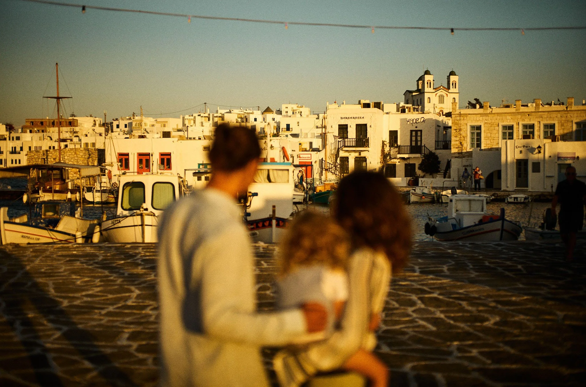 A blurred family of three standing on a cobblestone street near a harbor, with white buildings and boats in the background, during sunset.