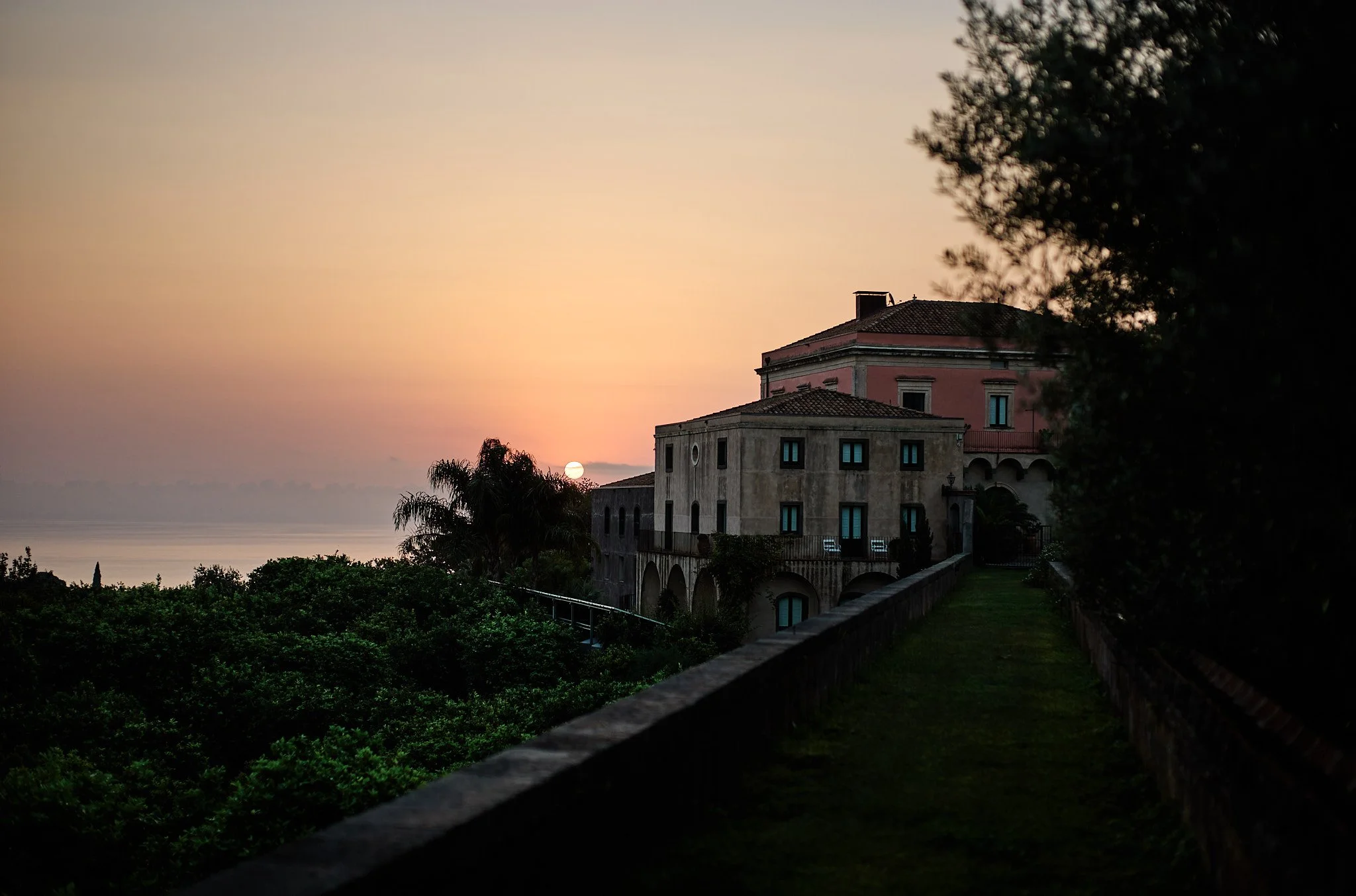 Sunset over a historic building with pink and orange sky, overlooking water with trees in the foreground.