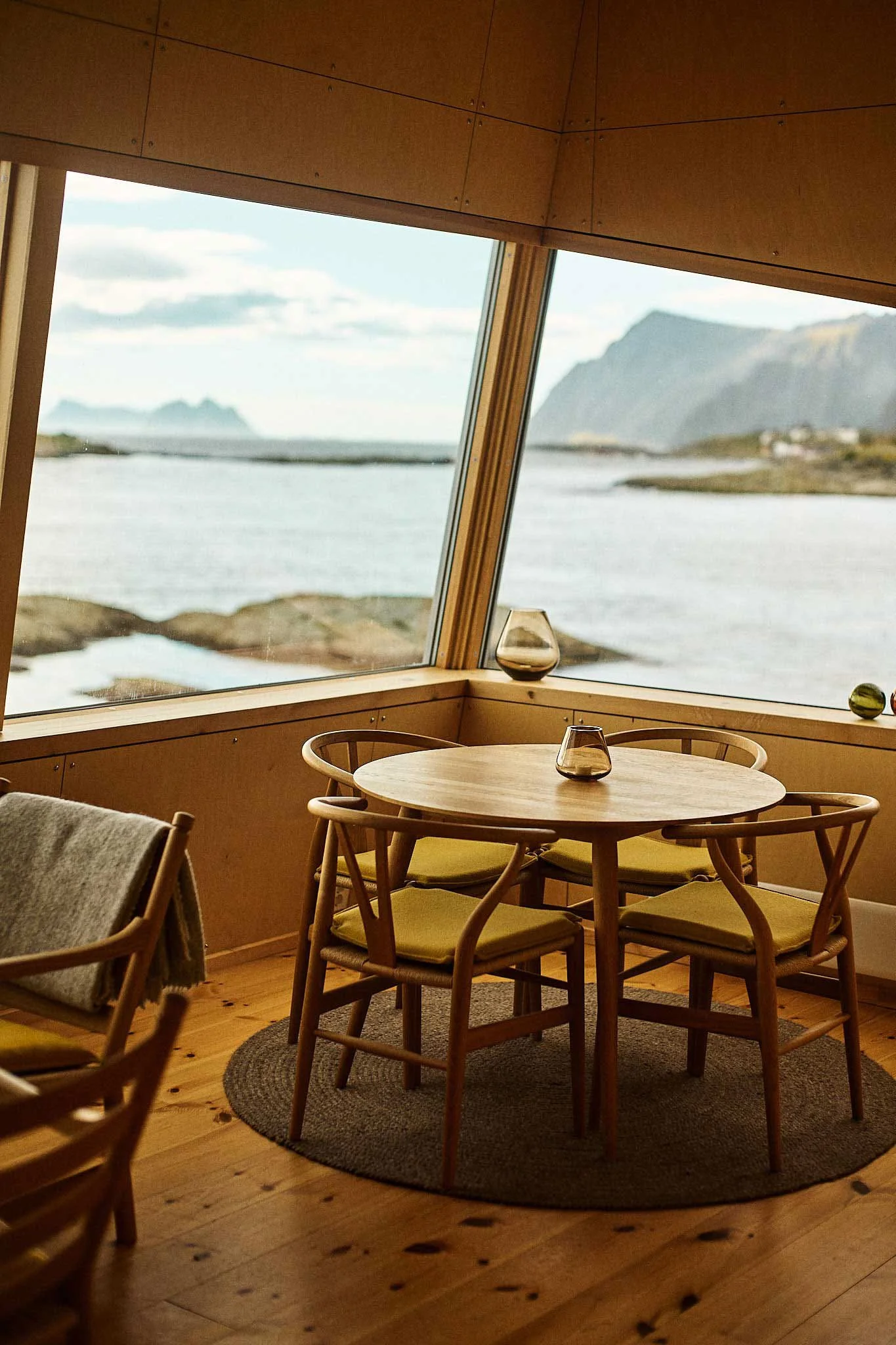 A cozy dining area with a round wooden table, four chairs with yellow cushions, and decorative glass vases on the window ledge. Large windows reveal a scenic view of water and mountains outside.