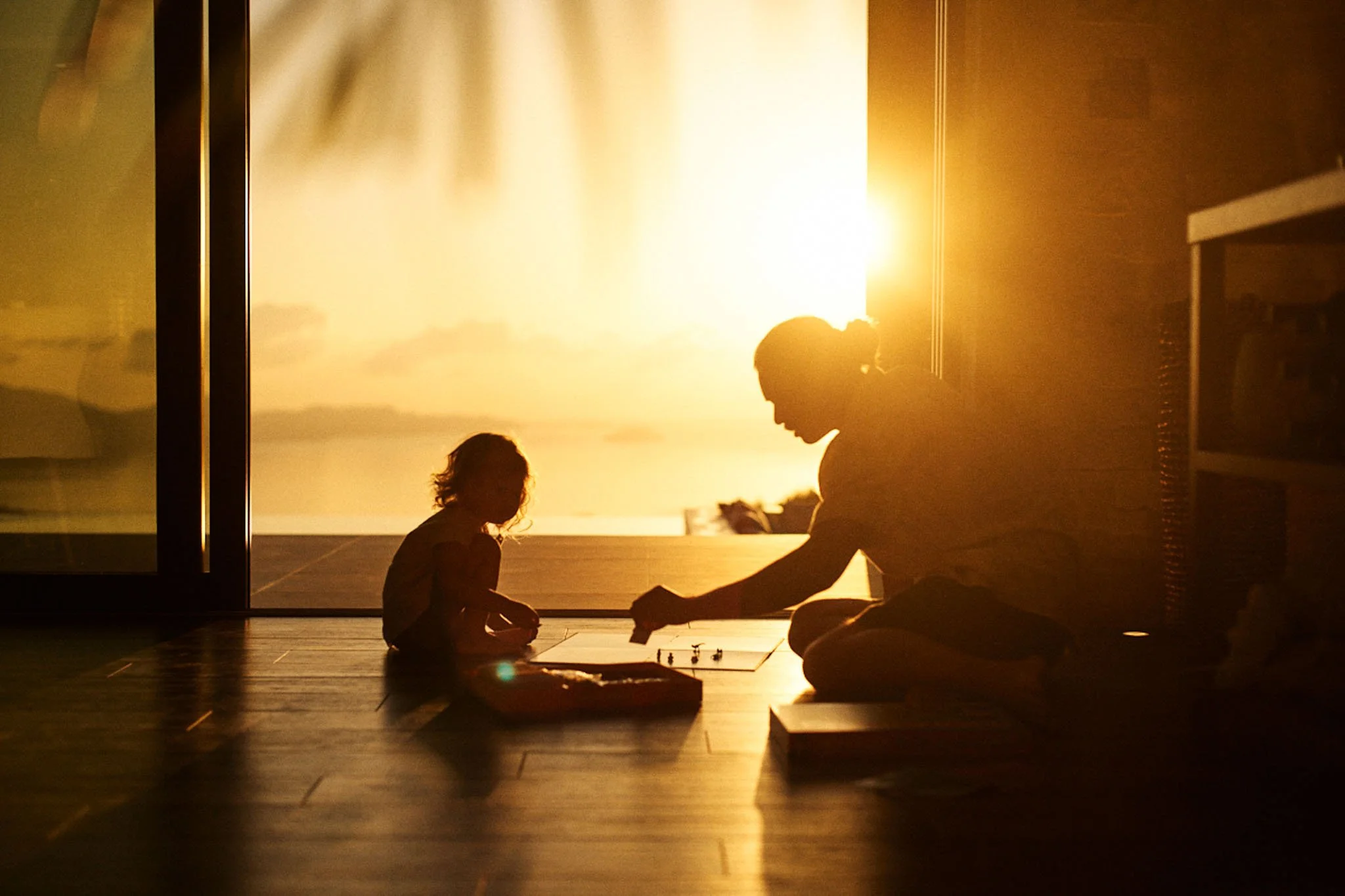 A woman and a young girl playing a board game on the floor at sunset, silhouette against a large window with a view of the beach and ocean.