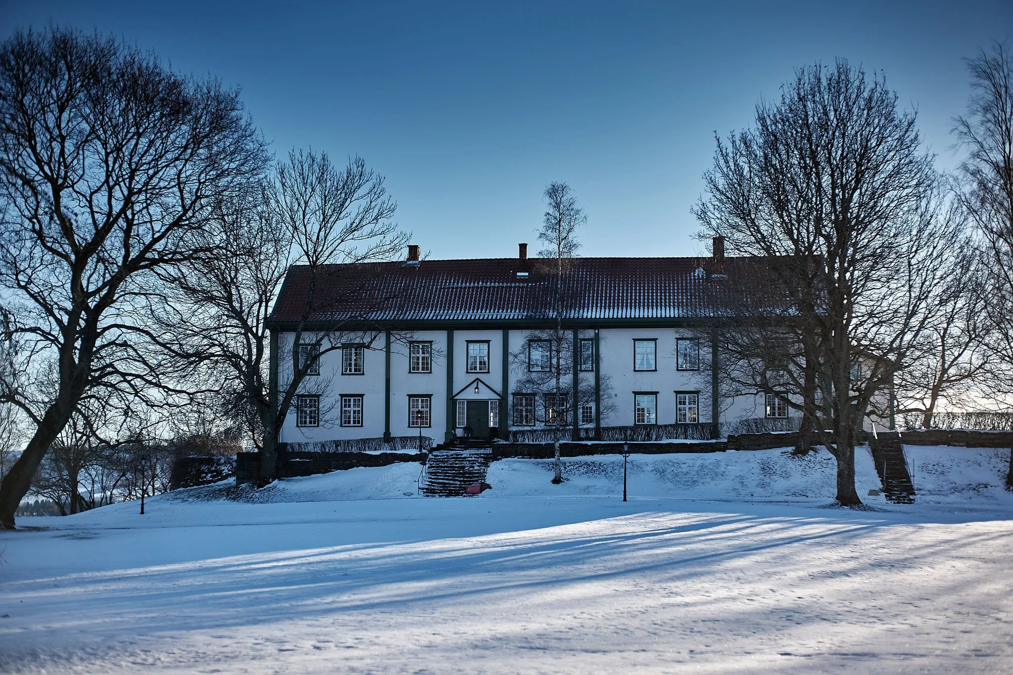 A large, white, two-story house with a red-tiled roof surrounded by snow and leafless trees, under a clear blue sky.