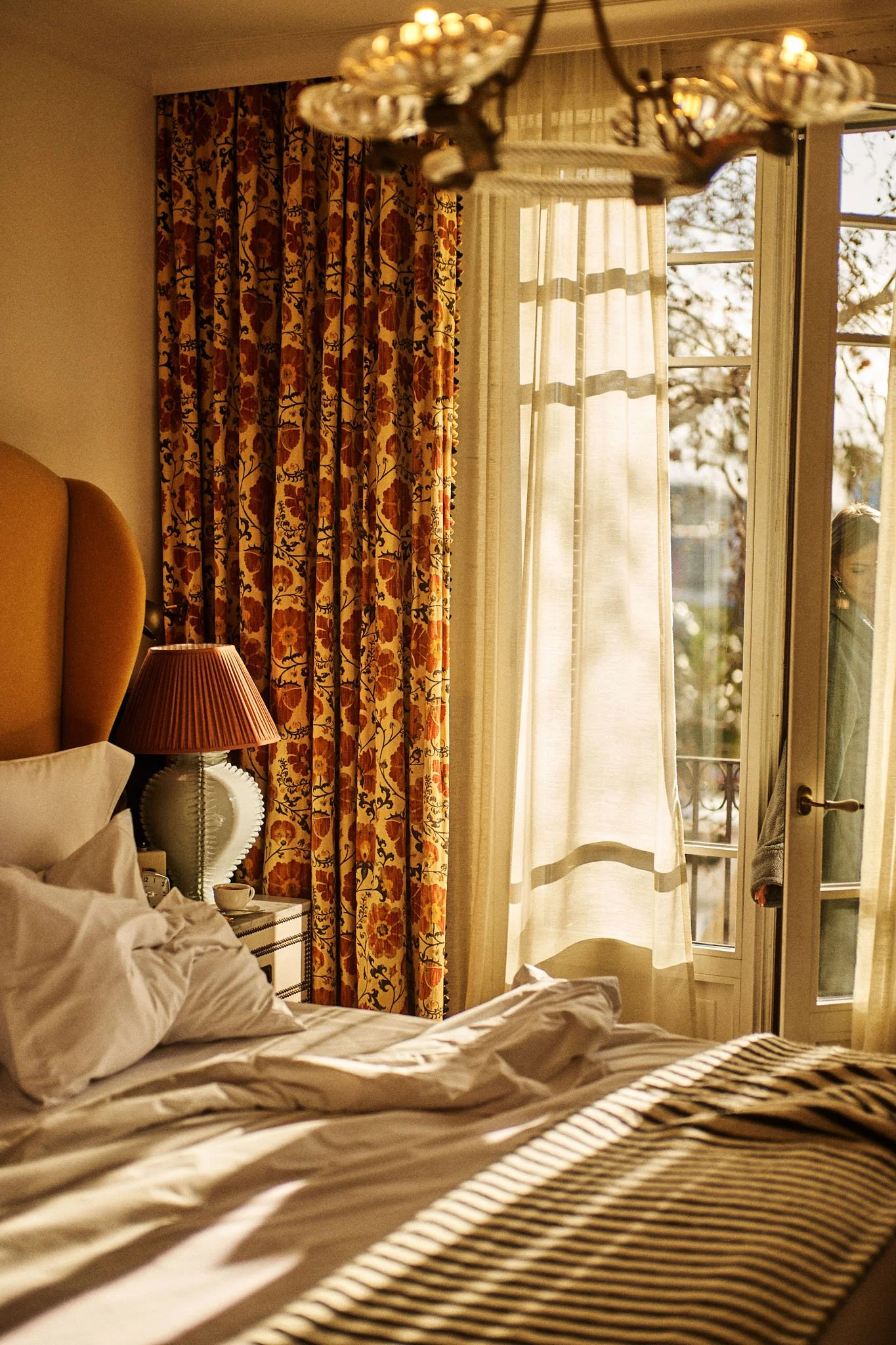 A cozy hotel room with an unmade bed, a nightstand with a lamp, and curtains partially open revealing a woman outside.