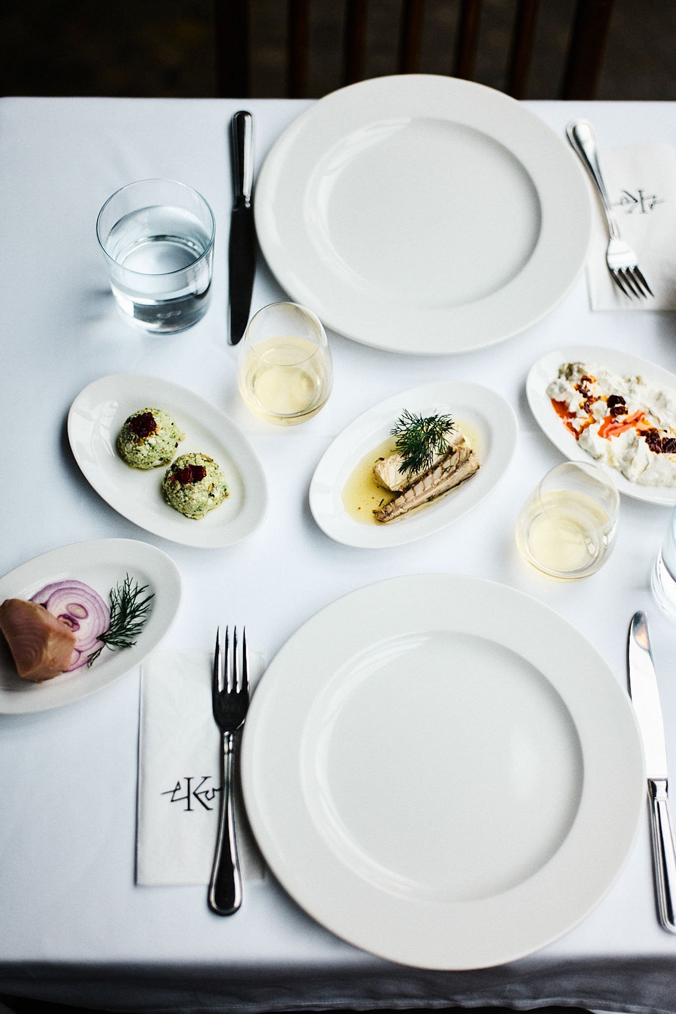 Empty white dinner plates with silverware, a glass of water, and a glass of white wine on a white tablecloth, with various appetizers including two rice balls, a slice of fish, a creamy dip with red garnish, and sliced onions with herbs.