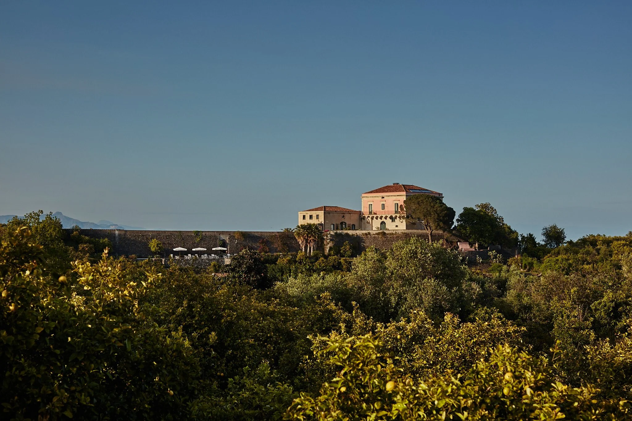 A castle-like building on a hilltop surrounded by trees and greenery with a clear blue sky in the background.