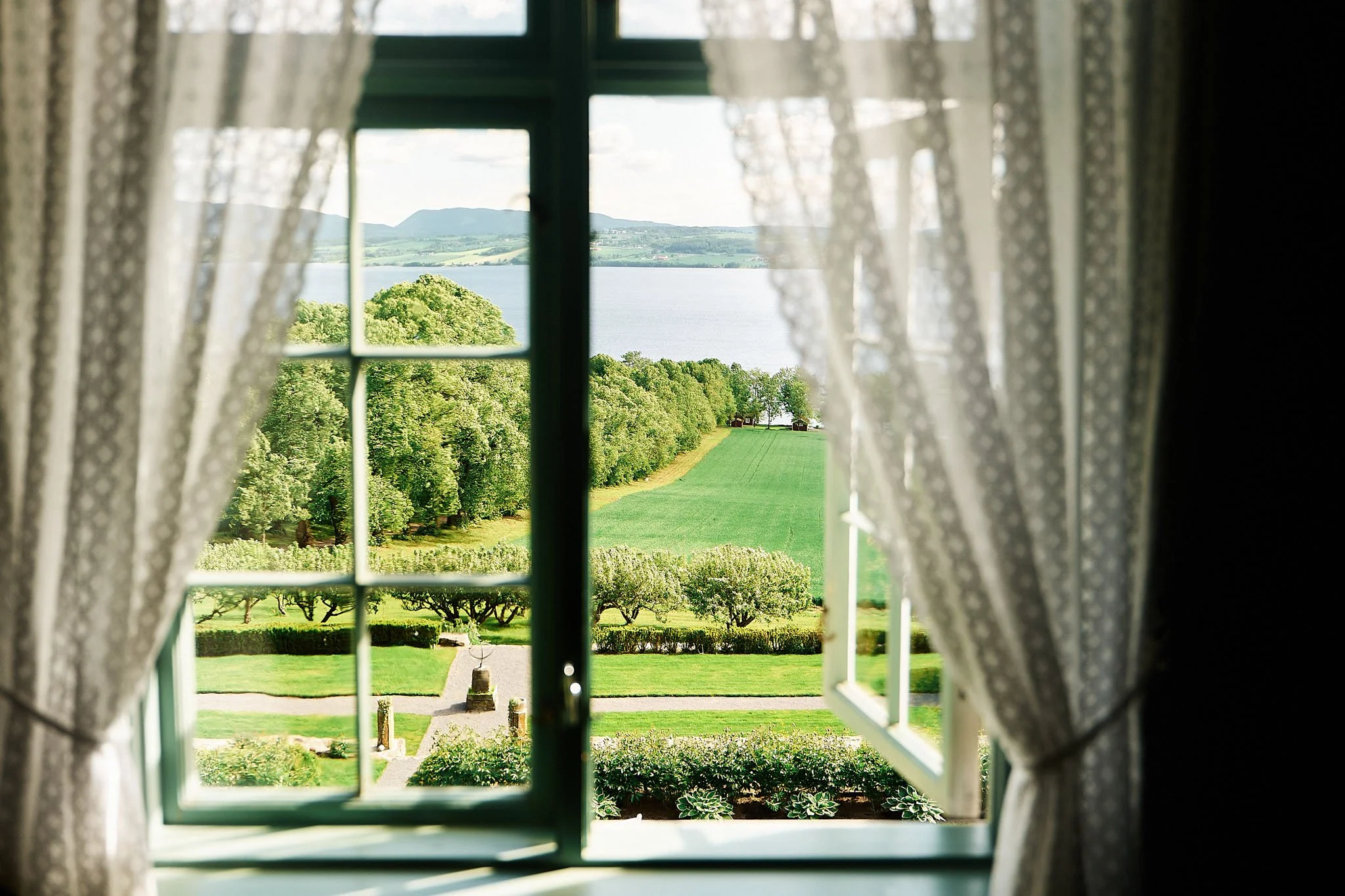 Scenic view of green trees, grass, water, and distant hills seen through an open window with curtains.