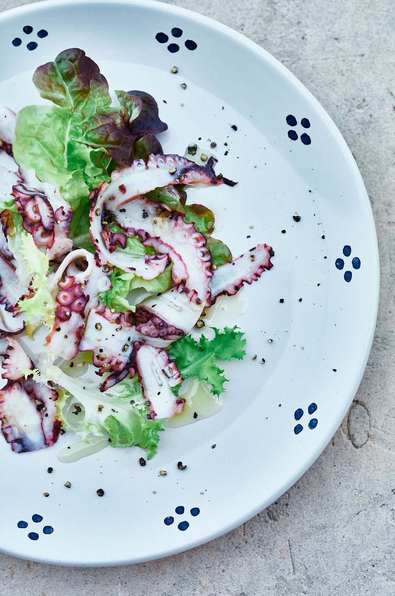 A plate of seafood salad with octopus, mixed greens, black pepper, and salt on a white plate with blue dots, placed on a gray surface.
