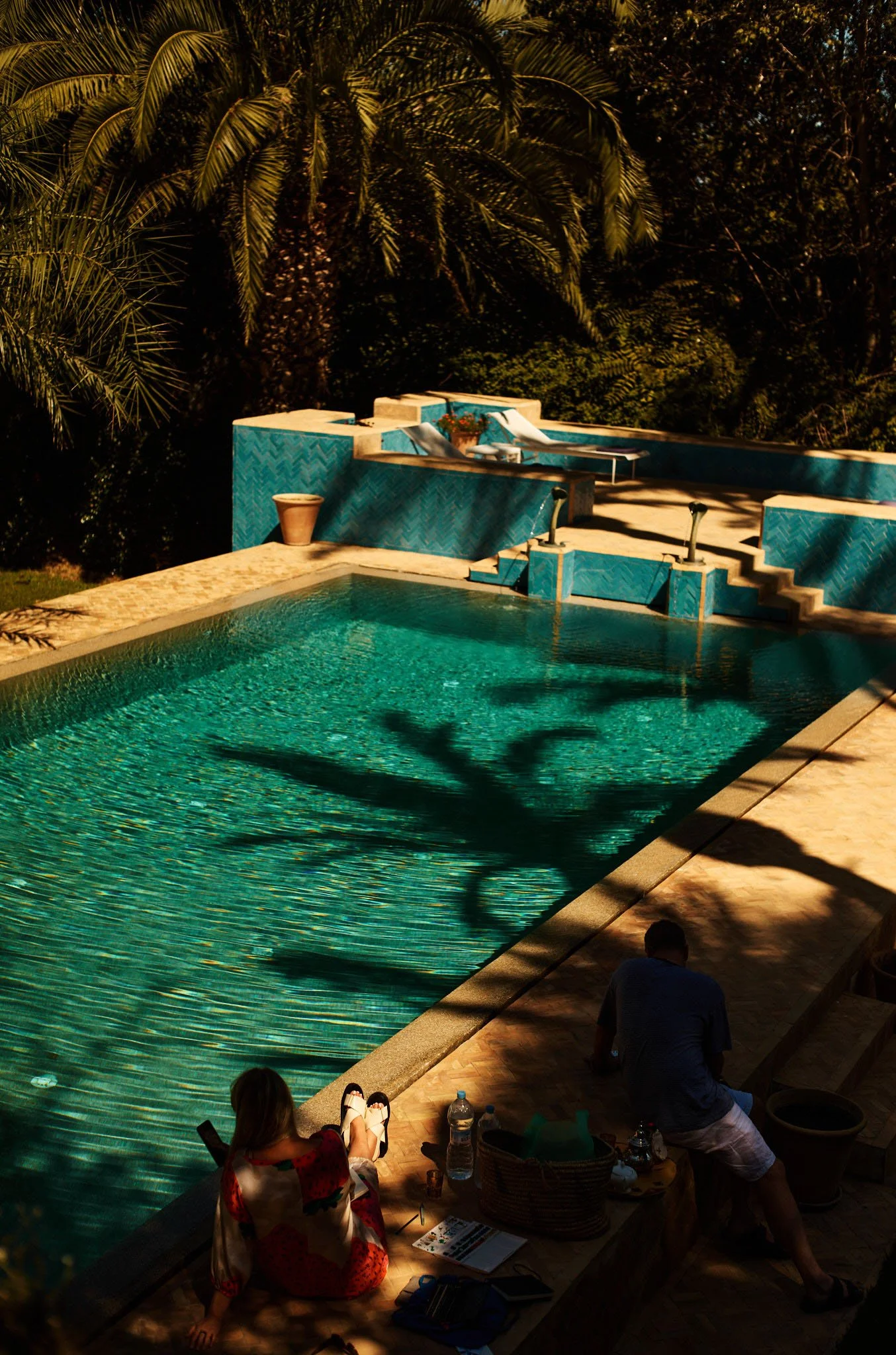 A swimming pool with blue tiles, surrounded by a beige stone deck. A woman in a colorful dress and a man are sitting by the poolside. Shadows of palm trees are cast on the ground and water, and lush tropical trees are in the background.