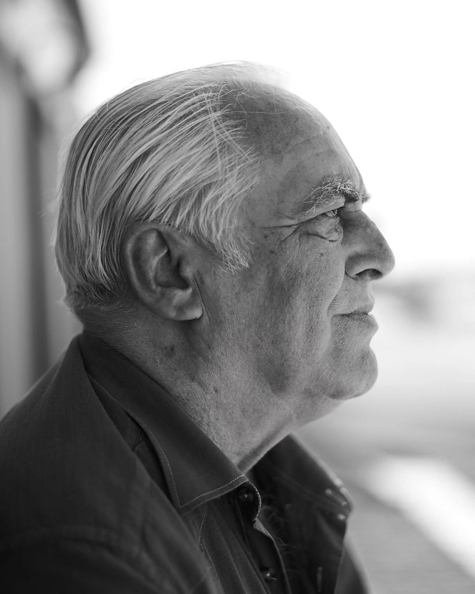 Profile view of an elderly man with white hair, wearing a dark collared shirt, looking pensively to the right.