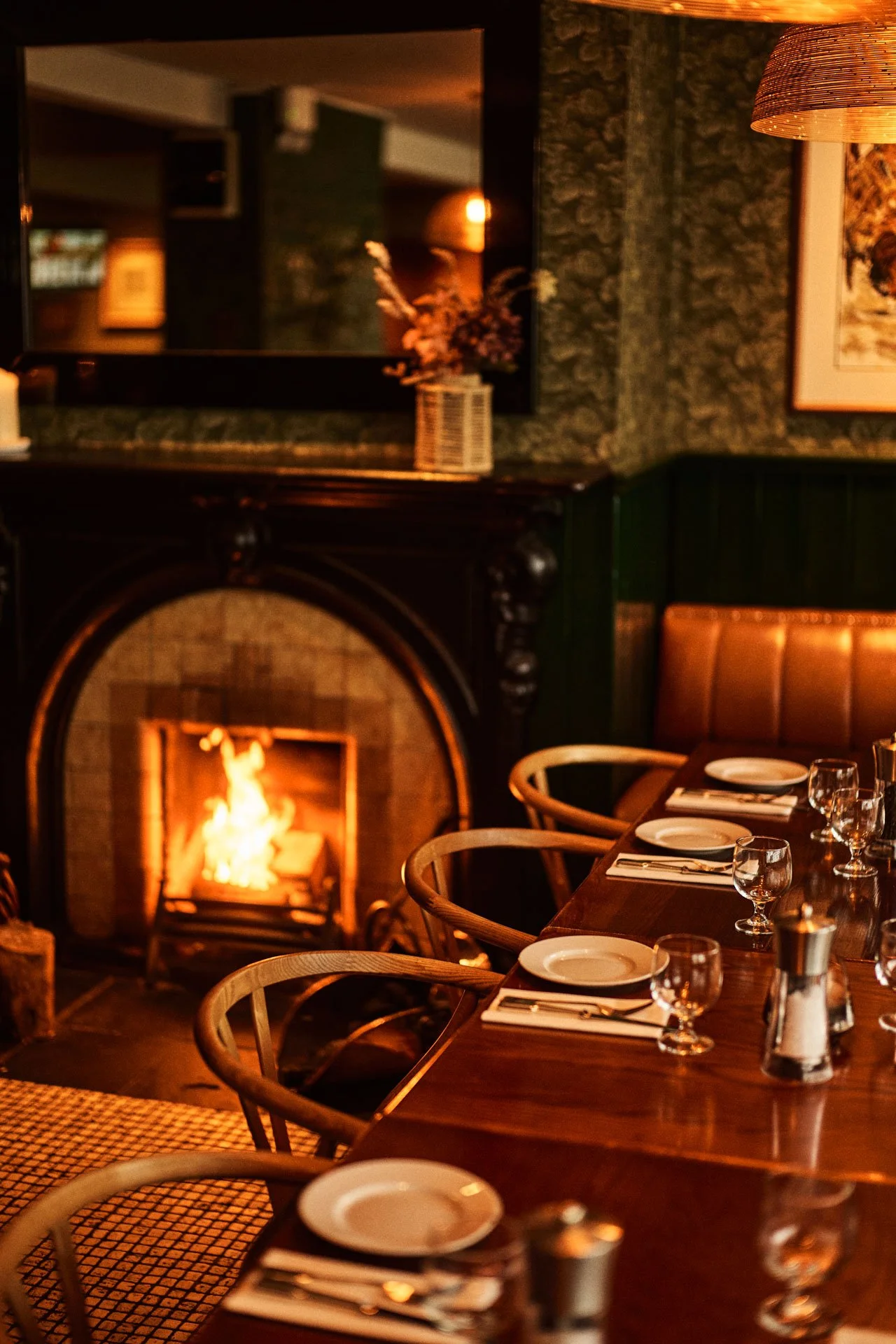 Cozy restaurant interior with a lit fireplace, a long wooden dining table set with plates, forks, knives, glasses, and napkins, and a mirror reflecting a floral arrangement and warm lighting.