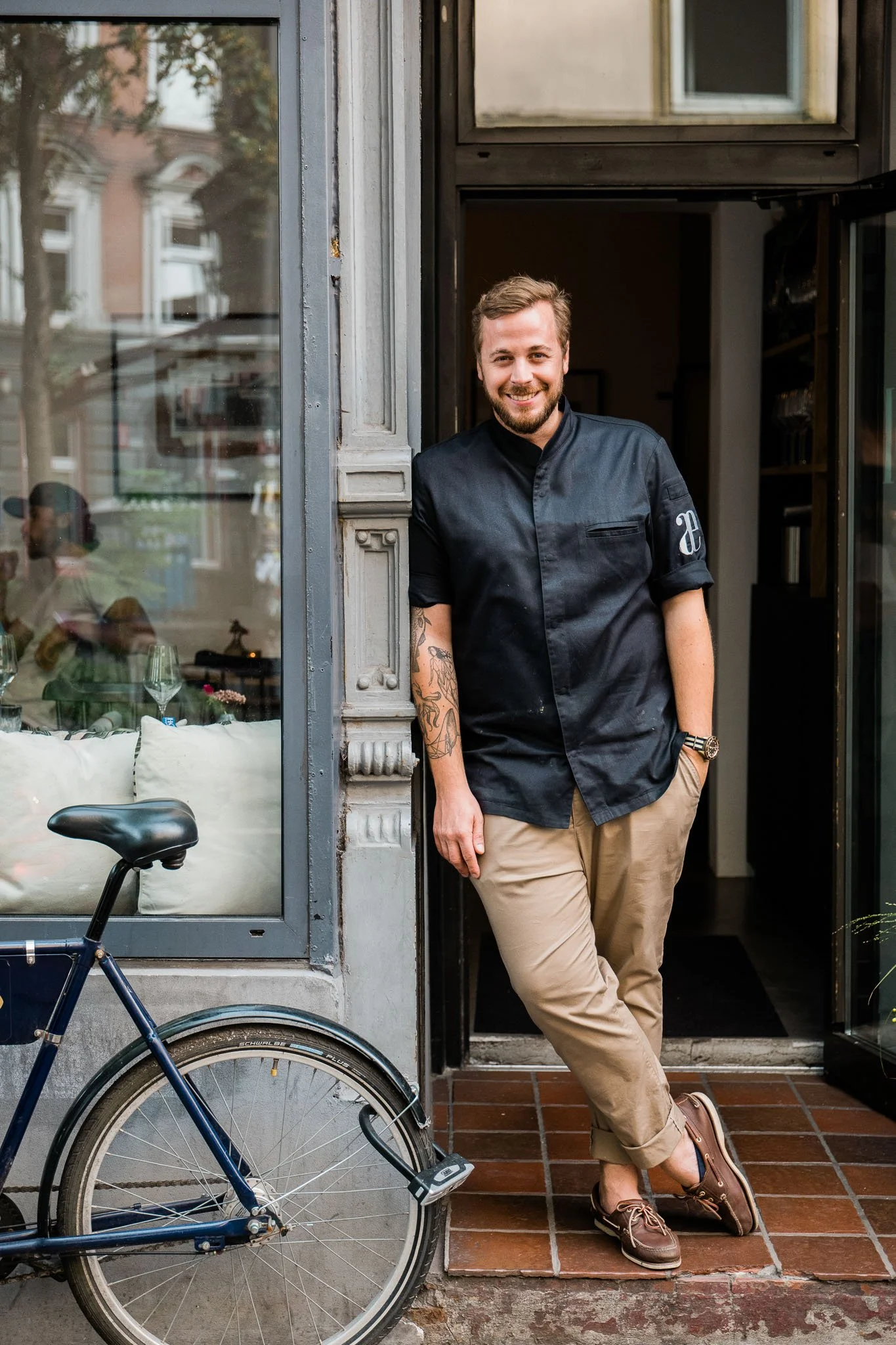 A smiling man with a beard, tattoos, and short hair standing at the entrance of a cafe or restaurant, wearing a black chef's jacket, beige pants, and brown shoes.