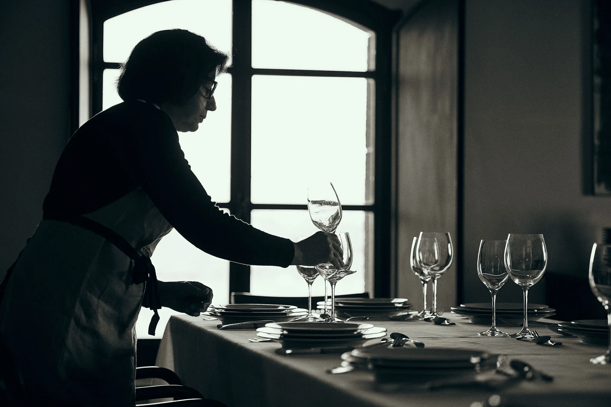Silhouette of a woman placing a wine glass on a dining table in a dimly lit room with large windows.