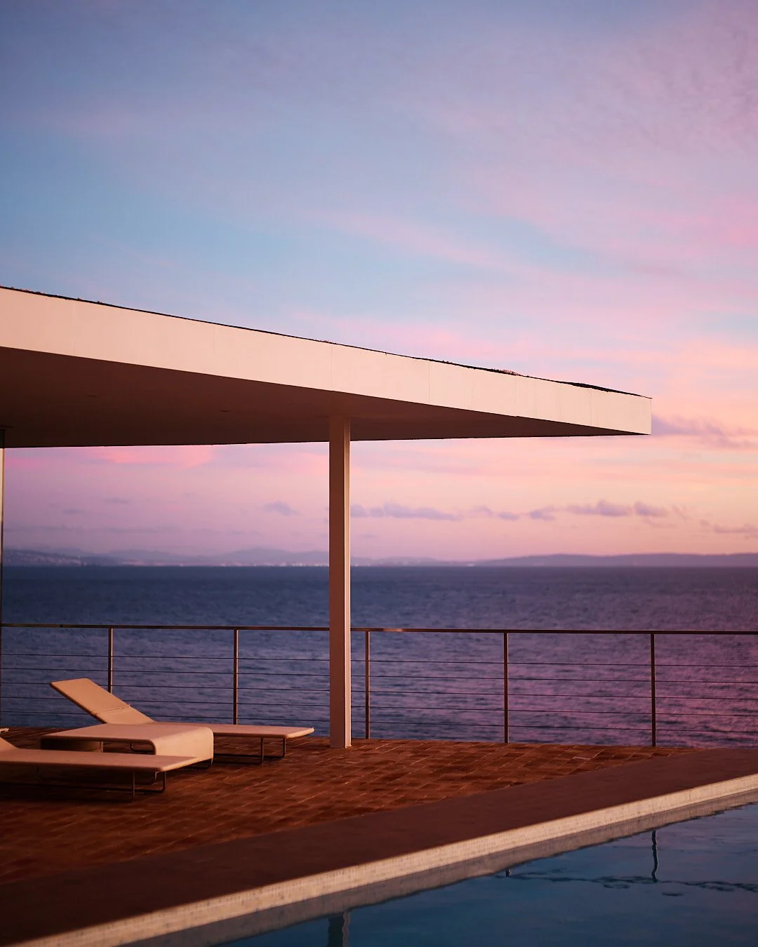 Sunset view from a swimming pool balcony overlooking the ocean with two lounge chairs and a modern white roof structure.